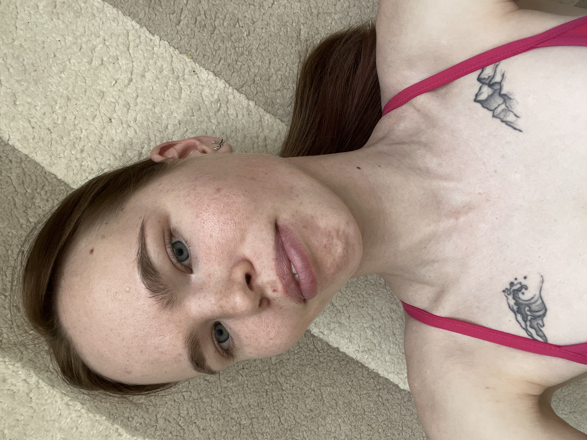 A young woman with fair skin, light blue eyes, and auburn hair is lying on a carpet of beige and white stripes. She is wearing a pink bikini top. Two dark grey tattoos are visible on her chest, one near each strap. The environment suggests a domestic interior. The location, Chaikovsky, Russia, is extraneous to the visual information present in the image.
