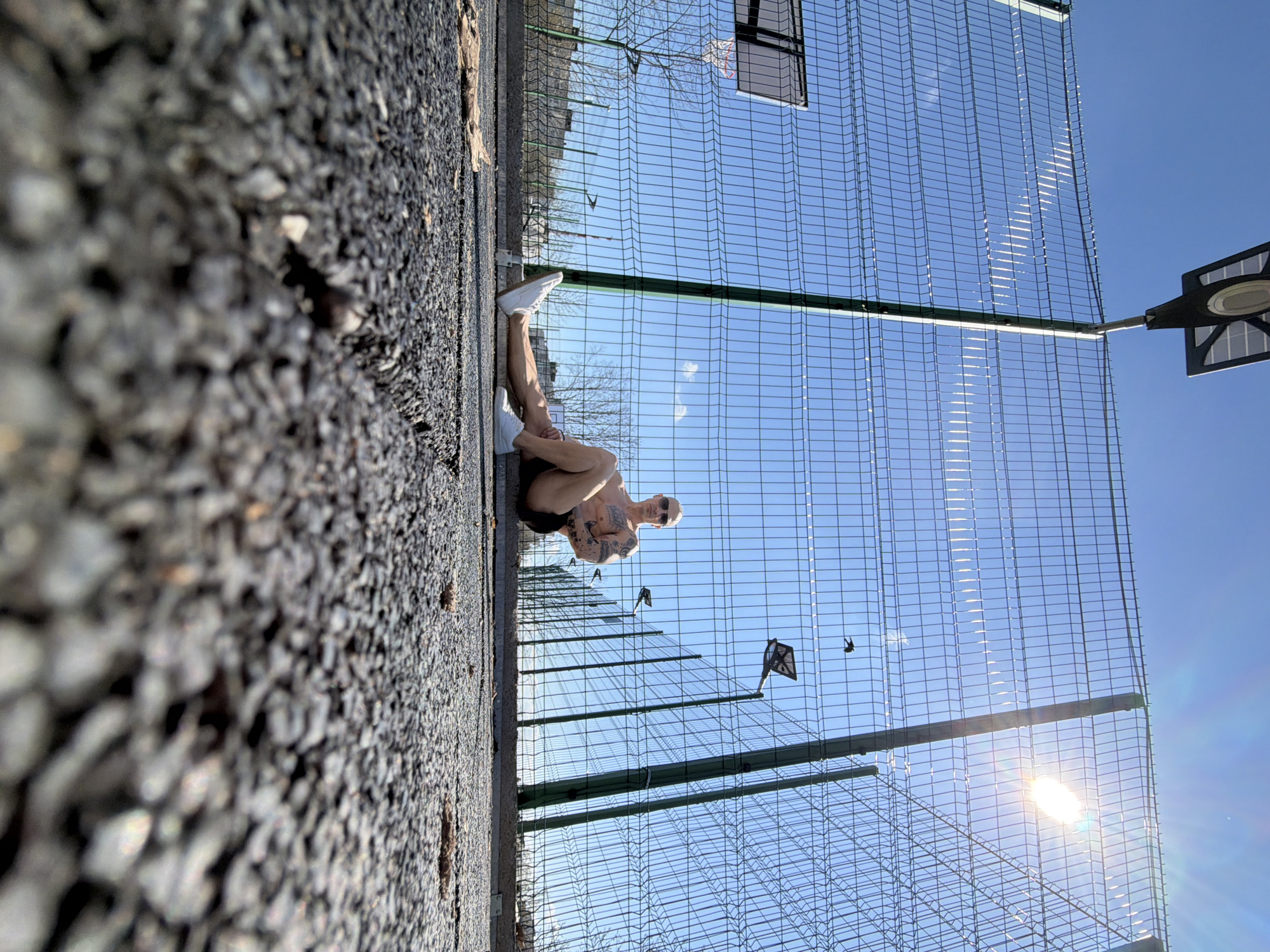 The image depicts a man sitting on a gravel surface next to a chain-link fence, in what appears to be an outdoor basketball or sports court. The man is wearing shorts, sneakers, and sunglasses, and he has tattoos on his arms. He is seated with his legs bent. The background consists of a blue sky and the fence of the sports court. Sunlight is visible through the fence. The image was taken in Pleven, Bulgaria.
