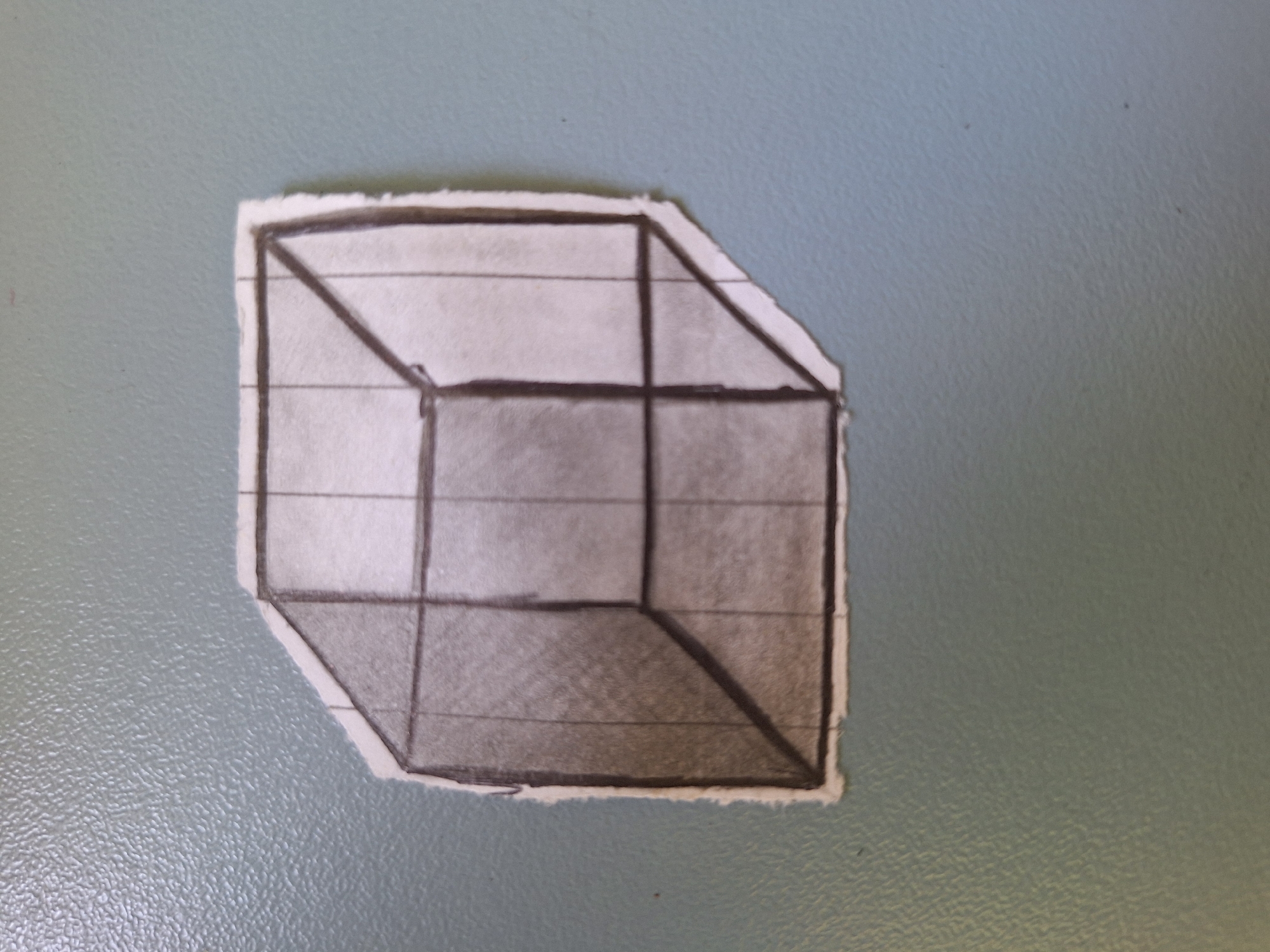 A torn piece of paper with a hand-drawn cube on it is affixed to a textured, light blue surface. The cube is drawn with thick, dark lines and filled with graphite shading to give it a three-dimensional appearance. The paper has uneven, torn edges. The background surface is consistent with a wall or cabinet. There are no people or animals present, and no context is provided to indicate a specific location in Varna, Bulgaria.