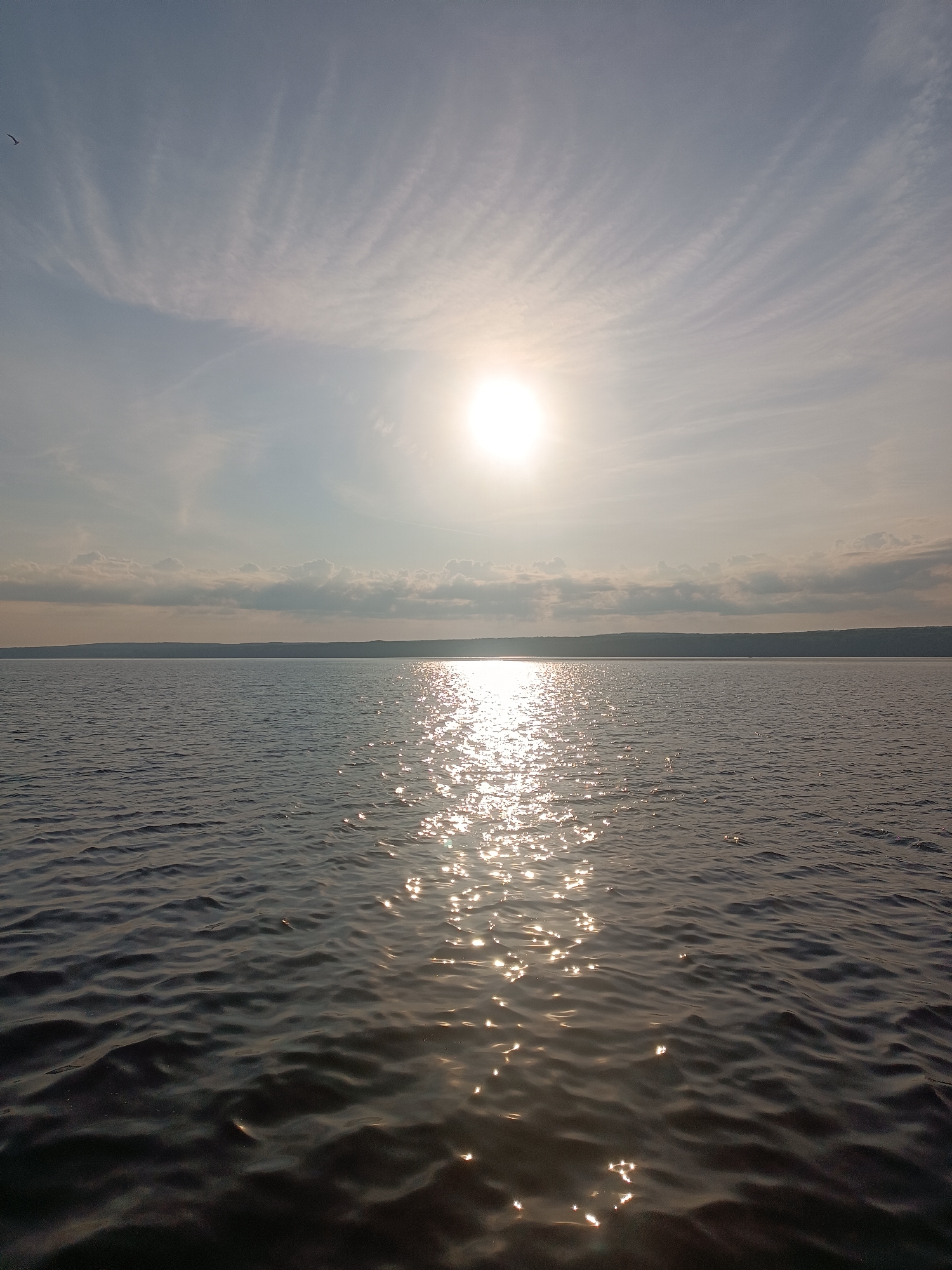 A wide shot captures a serene body of water, likely a river or lake, under a bright, hazy sun. The sun is positioned slightly off-center to the right in the upper portion of the frame, casting a shimmering reflection across the water's surface. The water itself is calm, with subtle ripples that catch the sunlight, creating a path of glittering light leading towards the viewer. In the distance, a flat, tree-lined horizon forms the edge of the land, with a soft line of clouds hugging the distant shore. The sky above is a pale blue, streaked with wispy, cirrus clouds that are illuminated by the sun. A small, dark bird is visible in flight in the upper left quadrant of the sky. The overall impression is one of tranquility and natural beauty, possibly taken from a boat or the shoreline. The location is Chaikovsky, Russia, specifically the Kama River, known for its wide expanse and scenic views.