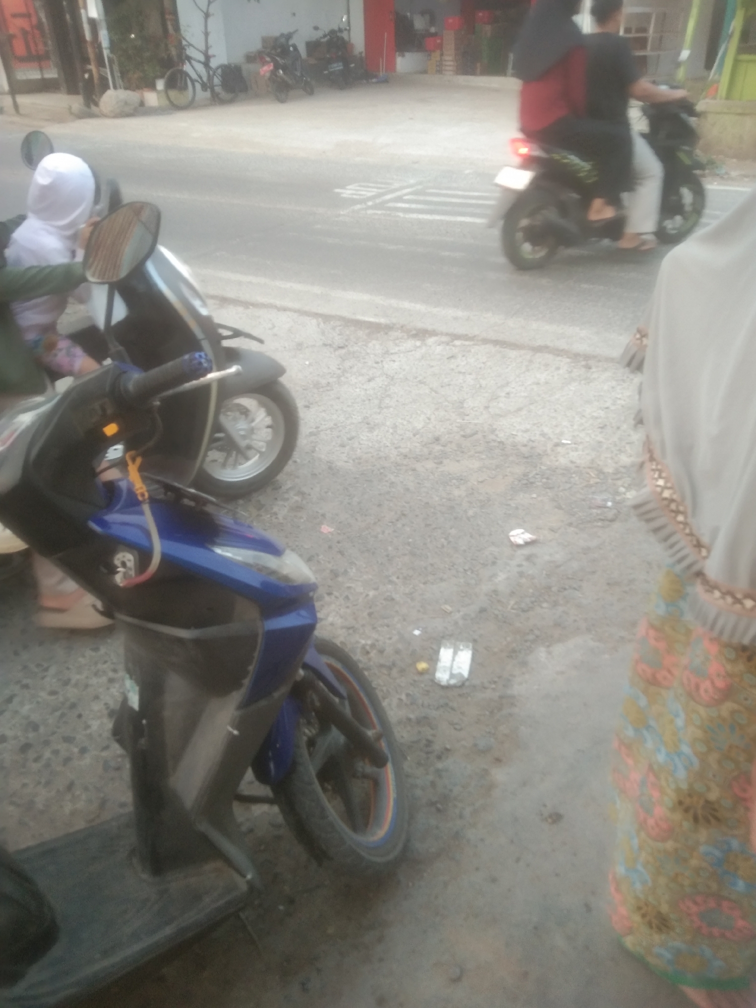 Two scooters are parked on a paved surface next to a road. The scooter in the foreground is blue and black, with a black floorboard and a single wheel visible. The scooter in the background is dark, with a silver wheel. Two individuals are on the background scooter; a person wearing a red shirt and black headscarf is driving, and another person wearing a black shirt and grey pants is a passenger. A person wearing a white headscarf is partially visible next to the foreground scooter. The environment appears to be a street in Subang, Indonesia, with buildings and trees visible in the background.