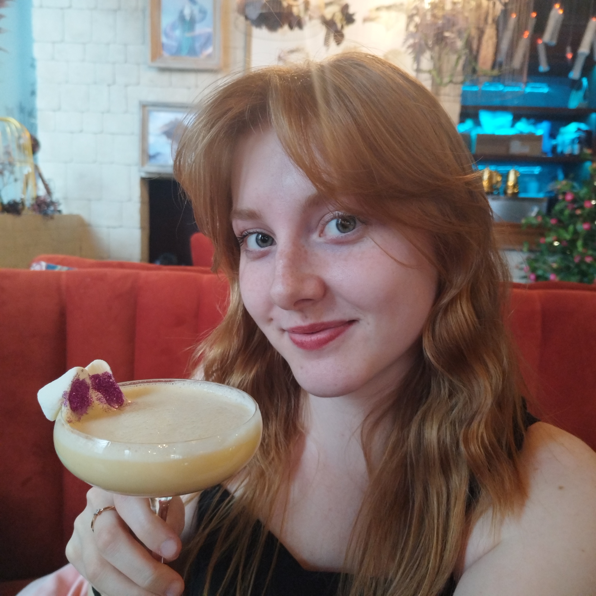 A young woman with long, wavy, reddish-brown hair, styled with bangs, is featured in a close-up, chest-up portrait. She has fair skin, light eyes, and a pleasant smile, showing a dimple on her left cheek. She wears a dark top and a delicate ring on her right ring finger. She holds a coupette glass containing a light yellow, frothy cocktail, which is garnished with two white, fluffy pieces, likely marshmallows, adorned with purple glitter.
The scene is an indoor setting, likely a bar or restaurant in Yekaterinburg, Russia. Behind her, there's a plush, red-orange upholstered booth or seating. The wall is made of white brick, with framed pictures visible further back. Various decorative elements include what appear to be dried flowers or ethereal hangings from the ceiling. To the right, a bar or shelving unit is illuminated with blue LED lights, and a small, festive plant-like decoration with pink flowers and lights is visible. The lighting suggests an indoor, possibly evening, ambiance. The woman appears happy and relaxed. No visible text is present.