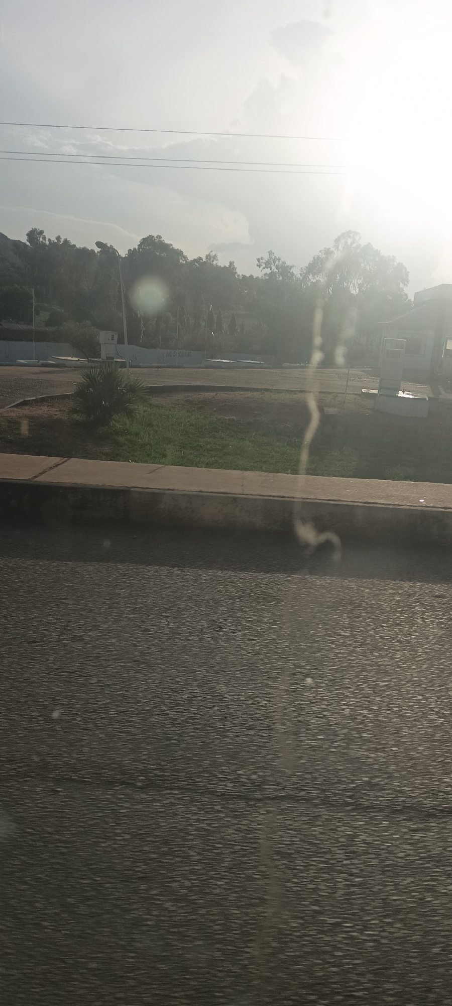 This media file captures an outdoor scene in Gura Top, Nigeria, viewed from what appears to be a moving vehicle. The image is significantly affected by bright sunlight and lens flare, particularly on the right side, creating a hazy, overexposed effect.
In the immediate foreground, a textured asphalt road is visible, exhibiting motion blur. Above the road, a concrete curb separates it from a grassy median strip with patches of dirt and small, green shrubs.
The midground features a long, white wall or sign on which the text "NO SMOKING" is clearly visible. To the right of this wall, there's a light-colored, small building or kiosk-like structure. A tall, dark utility pole stands in front of a dense line of trees that form a natural barrier.
The background consists of a prominent line of lush green trees, likely foliage on the slopes of hills, which are discernible in the distance on the left. Overhead power lines stretch across the upper part of the image against a bright, partly cloudy sky. The strong sunlight suggests either early morning or late afternoon.
No explicit activity or people are visible in the frame; the scene presents a static landscape observed from a dynamic perspective.