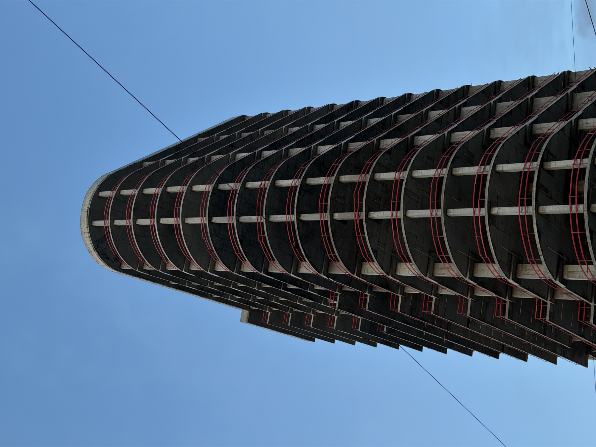 The image shows an unfinished high-rise building in Rostov-on-Don, Russia, seen from a low-angle perspective against a clear blue sky. The structure's facade consists of stacked, curving balconies supported by vertical red beams. The building's unfinished state reveals the bare concrete framework. Two dark cables are visible stretched diagonally across the upper portion of the frame.