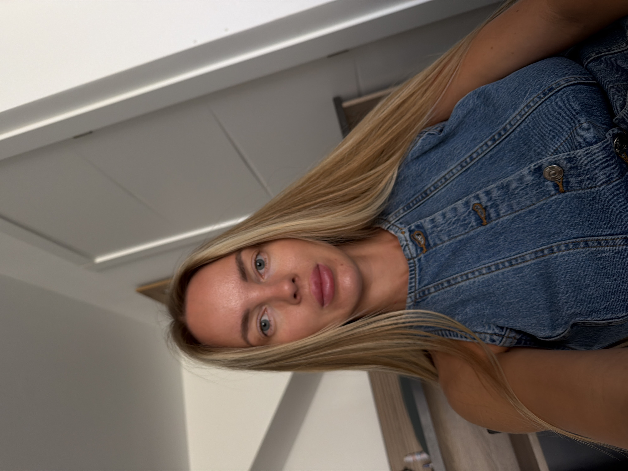 A woman with long blonde hair, wearing a denim vest, is seen from the chest up in a selfie. She is indoors, with a plain wall and a door visible in the background. The lighting suggests a natural light source. No specific location in Finestrat, Spain, can be identified from the image.