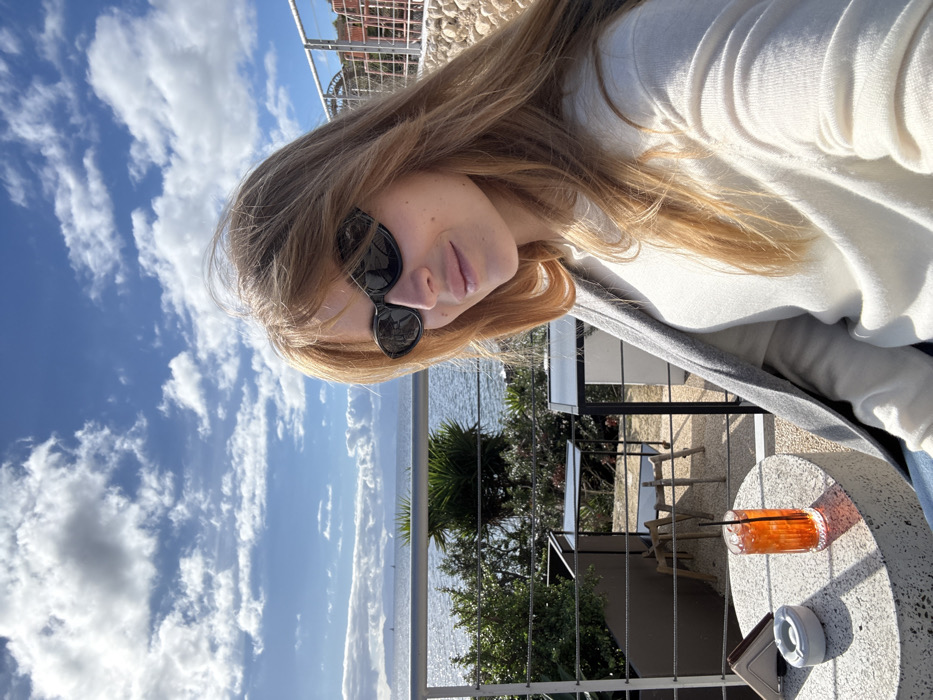 A female subject is captured in a half-body shot, seated at an outdoor terrace overlooking the Gulf of Cagliari in Cagliari, Italy. She occupies the right side of the frame, with her head and torso visible, smiling faintly towards the camera. Her long, light brown hair is parted centrally with bangs, and she wears dark, oval-shaped sunglasses. She is dressed in a light-colored (off-white) long-sleeved top with a grey cardigan draped over her shoulders.
In the lower left foreground, a round, textured concrete table displays an orange-hued beverage in a tall glass with ice and a dark straw, a white ashtray, and a stack of brown napkins. Behind the subject and table, a horizontal metal railing serves as a barrier for the terrace. Beyond this railing, green foliage, including bushes and a palm-like plant, is visible, preceding a lower-level outdoor seating area featuring several empty black-framed tables with glass tops and wooden chairs.
The background reveals the expanse of the blue Mediterranean Sea (Gulf of Cagliari) under a partially cloudy sky with prominent white cumulus clouds. Distant landmasses, appearing as hills and mountains, stretch across the horizon. Two small sailboats are discernible on the water's surface. The scene is illuminated by bright daylight, indicating clear weather.