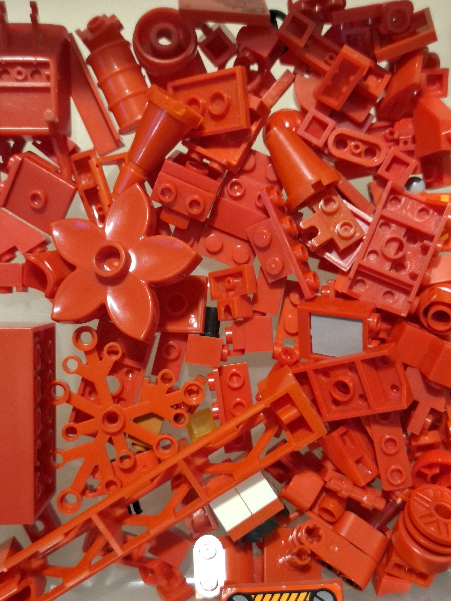 The image displays a collection of red plastic building blocks, possibly LEGOs or a similar toy. The blocks are arranged in a pile, obscuring much of the surface beneath. Various shapes and sizes of blocks are visible, including bricks, plates, specialized pieces like a flower-shaped piece, and pieces with wheels and connectors. The lighting appears to be from above, casting shadows within the pile. There is no discernible location in the city of Tyumen, Russia, or any people, actions, or interactions present in the image.
