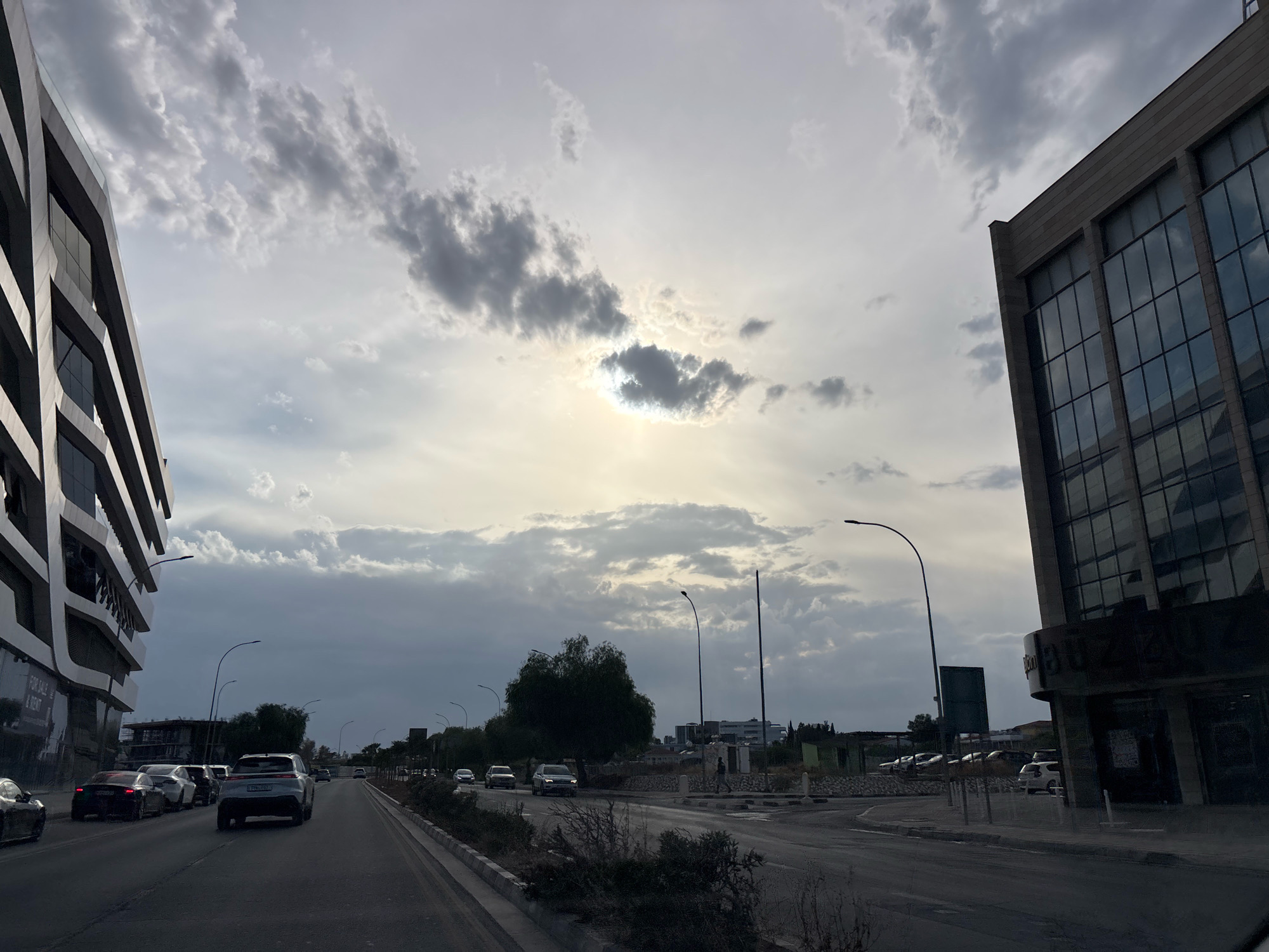This is a street-level photograph taken from a vehicle, looking forward and slightly to the right, in Mesa Geitonia, Cyprus. The sky is overcast with dramatic, textured clouds, with the sun peeking through in the upper center, creating a bright halo effect. On the left side of the road is a modern, multi-story building with a dark, angular facade and numerous reflective windows. A "For Sale & Rent" sign is visible on the lower part of this building. On the right side of the road is another large, dark building with a grid of reflective windows. The road itself is paved and appears to be a multi-lane street with a concrete curb and sidewalk. Several cars are on the road, including a white SUV in the foreground and other vehicles behind it. There are streetlights visible along the roadside. Trees and more distant buildings are visible in the background, suggesting an urban or suburban environment. The overall scene depicts a street view during what appears to be either early morning or late afternoon due to the lighting conditions.