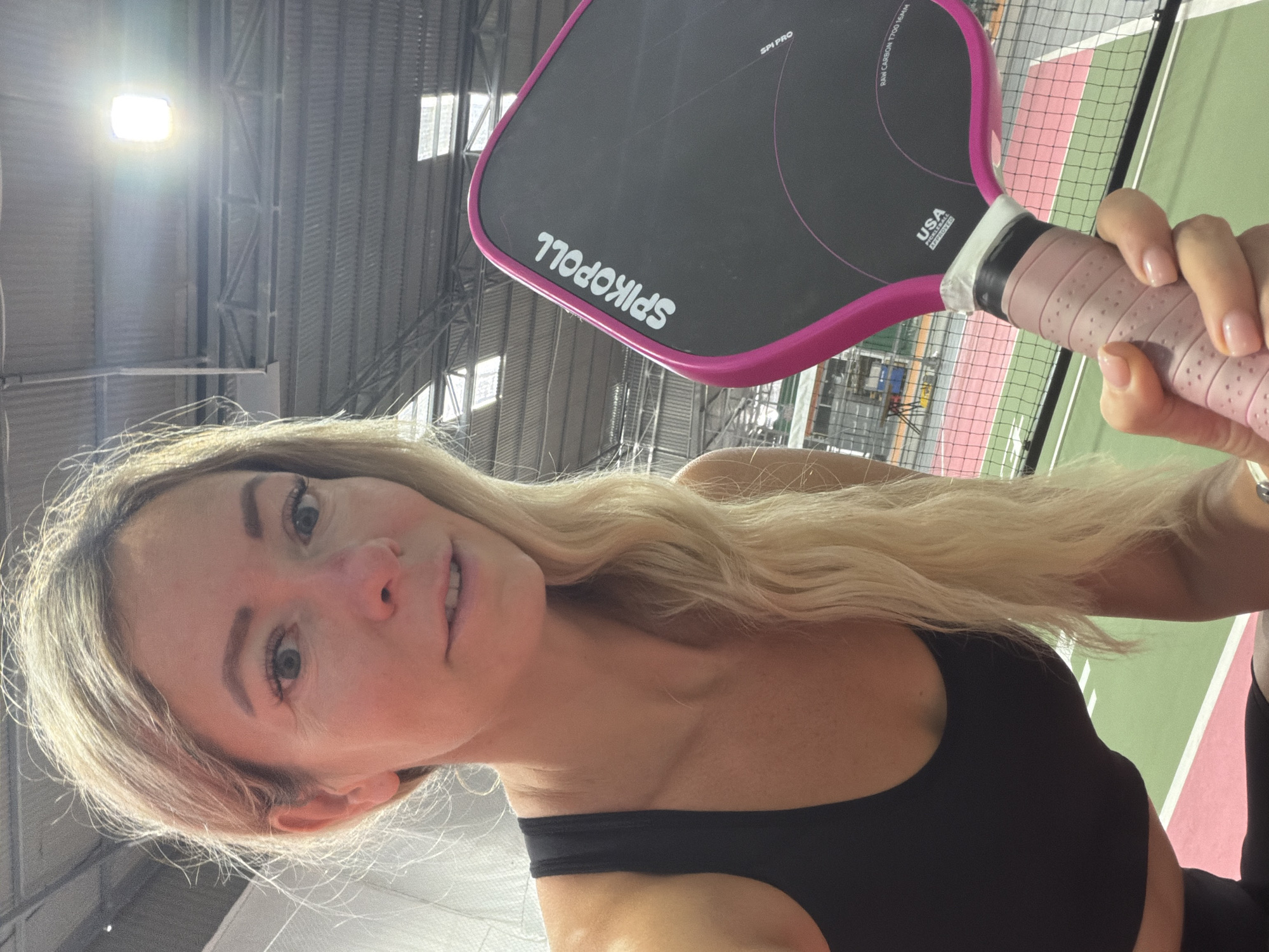 An indoor pickleball court with a woman holding a pickleball paddle. The woman has blonde hair and is wearing a black sports bra. The paddle is pink and black with the brand name "SPIKOPOLL" visible. The court has green and pink surfacing with a net visible in the background. The ceiling is industrial with visible trusses and lighting. There is no indication of the city of Da Lat, Vietnam in the image.