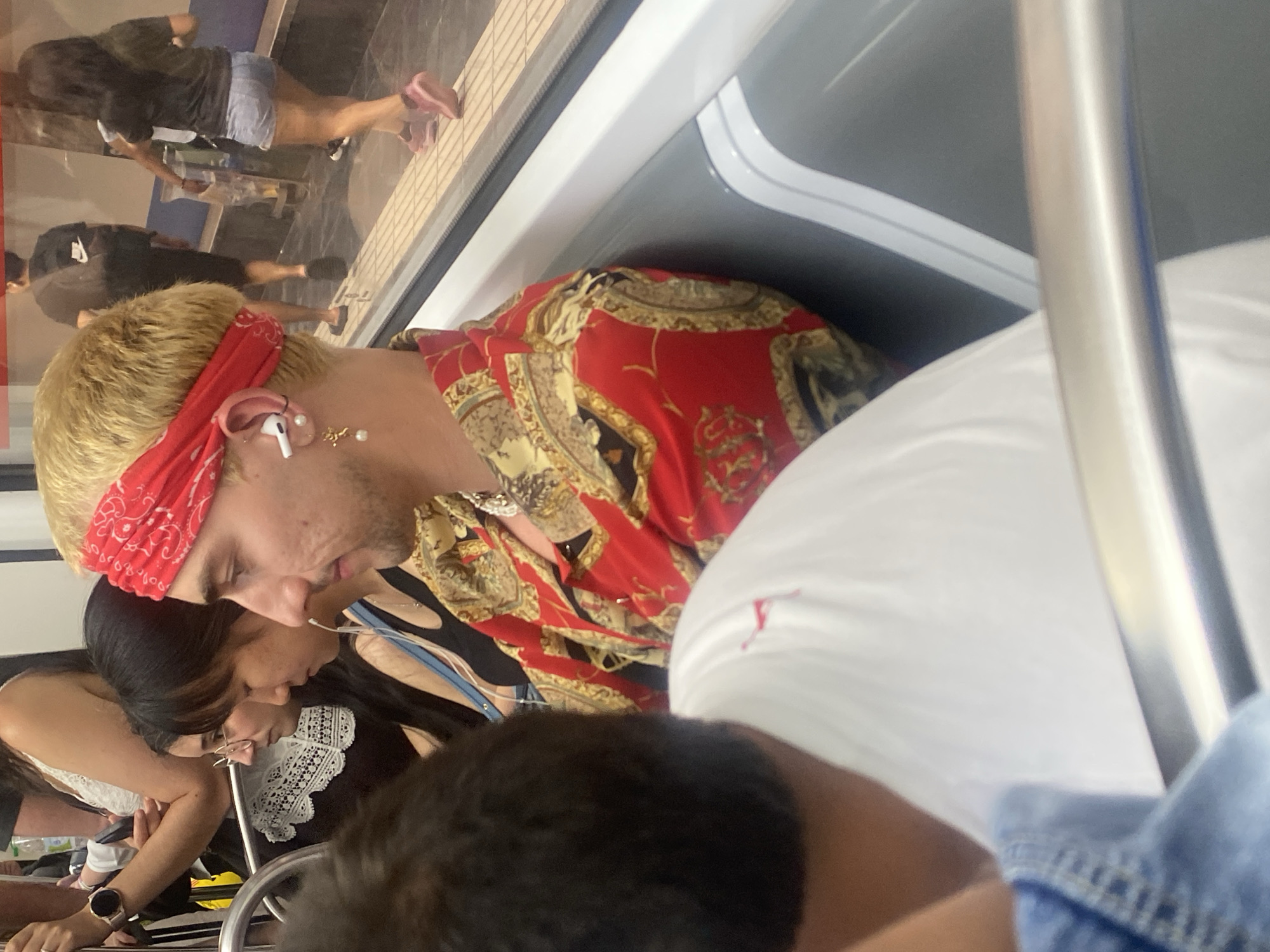The image captures a candid moment inside a public transportation vehicle, likely a subway or train, in L'Hospitalet de Llobregat, Spain. The focus is on a young man with dyed blonde hair, wearing a red bandana and a vibrant, gold-patterned red shirt. He has an AirPod in his ear and a pearl earring. He is seated next to two women; one is wearing glasses and a white top with lace detailing, and the other is wearing a black top with a blue strap. Behind them, other passengers are visible, including a person in a black t-shirt and shorts with a backpack, and a woman wearing a dark top and denim shorts. The interior of the vehicle features white seats and a metal railing, with a window revealing the station platform and other individuals. The overall atmosphere is casual and everyday, depicting a slice of urban life.