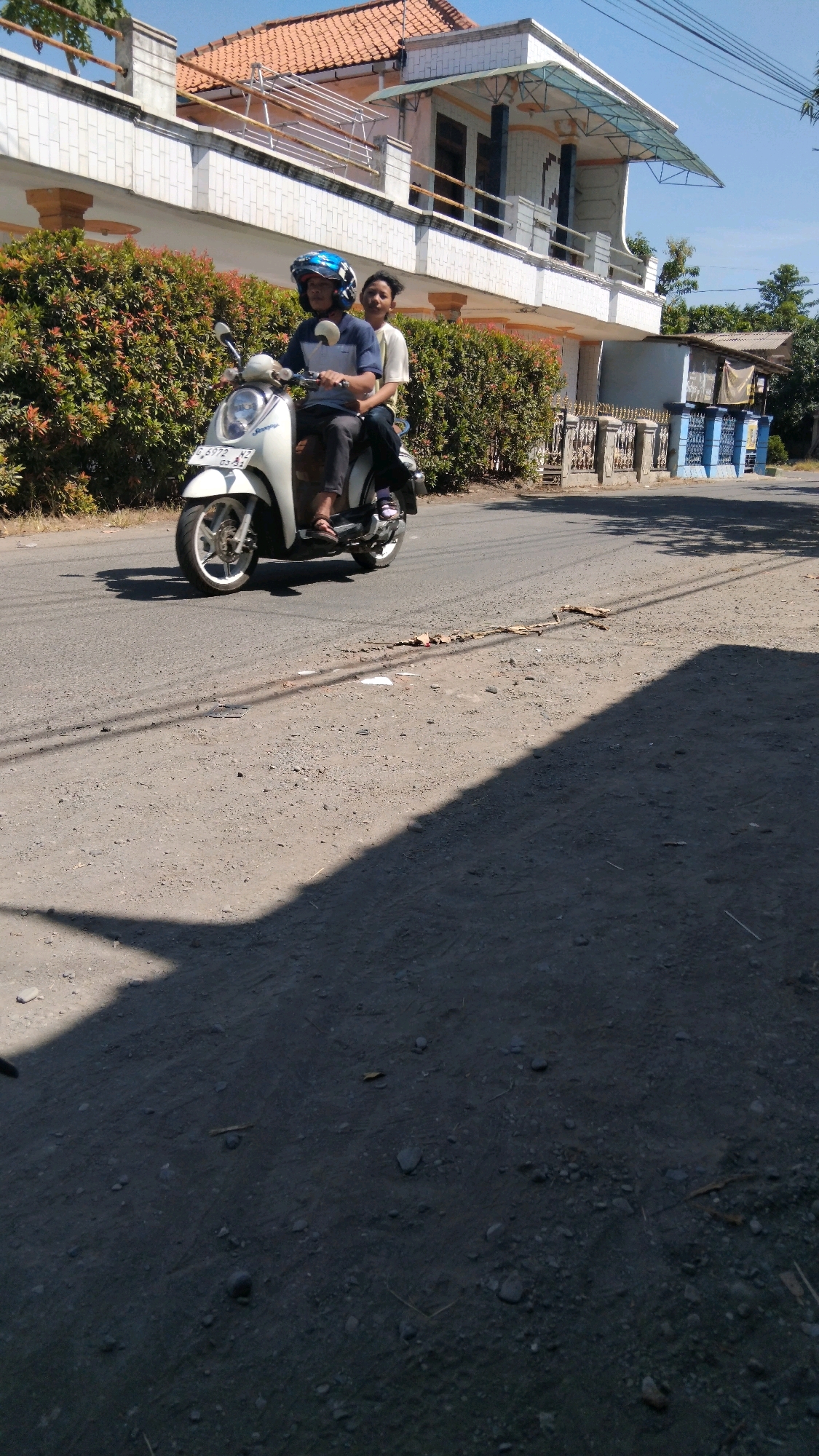 In Sidakaton, Indonesia, on a bright, sunny day, two individuals ride a white scooter down a paved road. The driver, wearing a blue helmet, navigates the scooter while the passenger sits behind him. The scooter is a modern model, suggesting it is likely a common mode of transportation in the area. The road is flanked by lush green bushes and a white house with a tiled roof, indicating a residential or suburban setting. The overall scene captures a slice of everyday life in this Indonesian town.