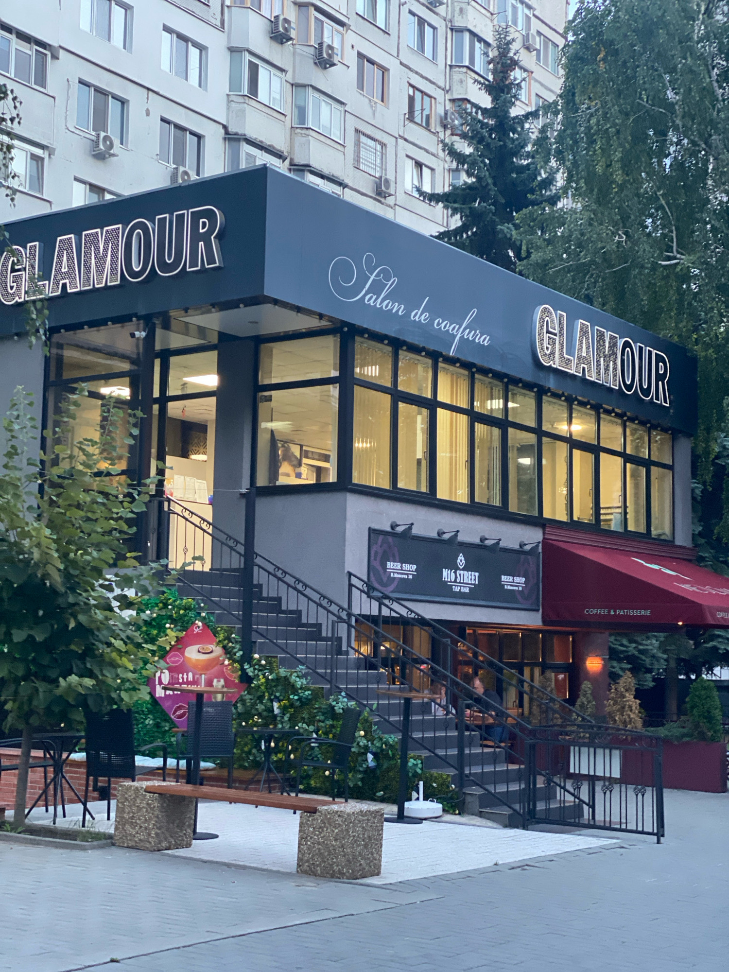 The image depicts the exterior of a building in Chișinău, Moldova, which houses multiple establishments. The most prominent feature is the signage for "GLAMOUR" in large, illuminated letters above the second floor, accompanied by "Salon de coafura" in elegant script. The building has a modern facade with large windows on the second floor, reflecting the interior lighting. Below the "GLAMOUR" sign, there is another level with signage for "BEER SHOP", "M16 STREET TAP BAR", and "COFFEE & PATISSERIE". A red awning extends from the "COFFEE & PATISSERIE" section.
A grey staircase with black metal railings leads up to the entrance on the second floor. Christmas decorations, including artificial trees and lights, are visible at the base of the stairs and around the entrance area, suggesting a festive season. In front of the building, there is a paved area with outdoor seating, including tables and chairs, and a concrete bench with a wooden seat. A tree is situated to the left of the entrance. The background shows a multi-story residential building with numerous windows and air conditioning units, and more trees. The overall impression is a commercial establishment in a mixed-use urban environment.