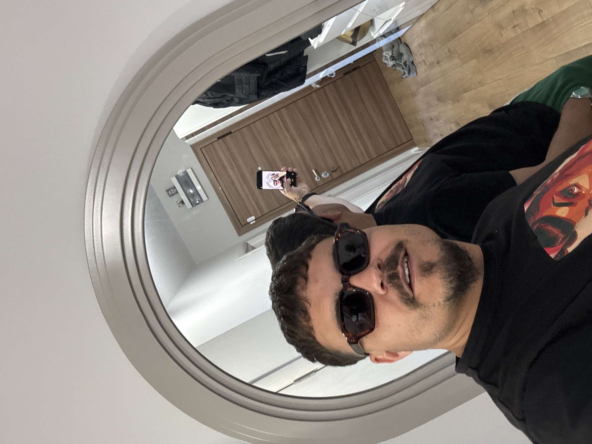 The image shows a selfie taken in a mirror. A man with dark hair, a goatee, and sunglasses is looking at the camera. He is wearing a black t-shirt. His reflection is visible in the mirror, showing him holding a phone. The reflection of the room can be seen in the mirror, including a wooden cabinet, a wall with a picture frame, and wooden flooring. The location is Unknown, Unknown.