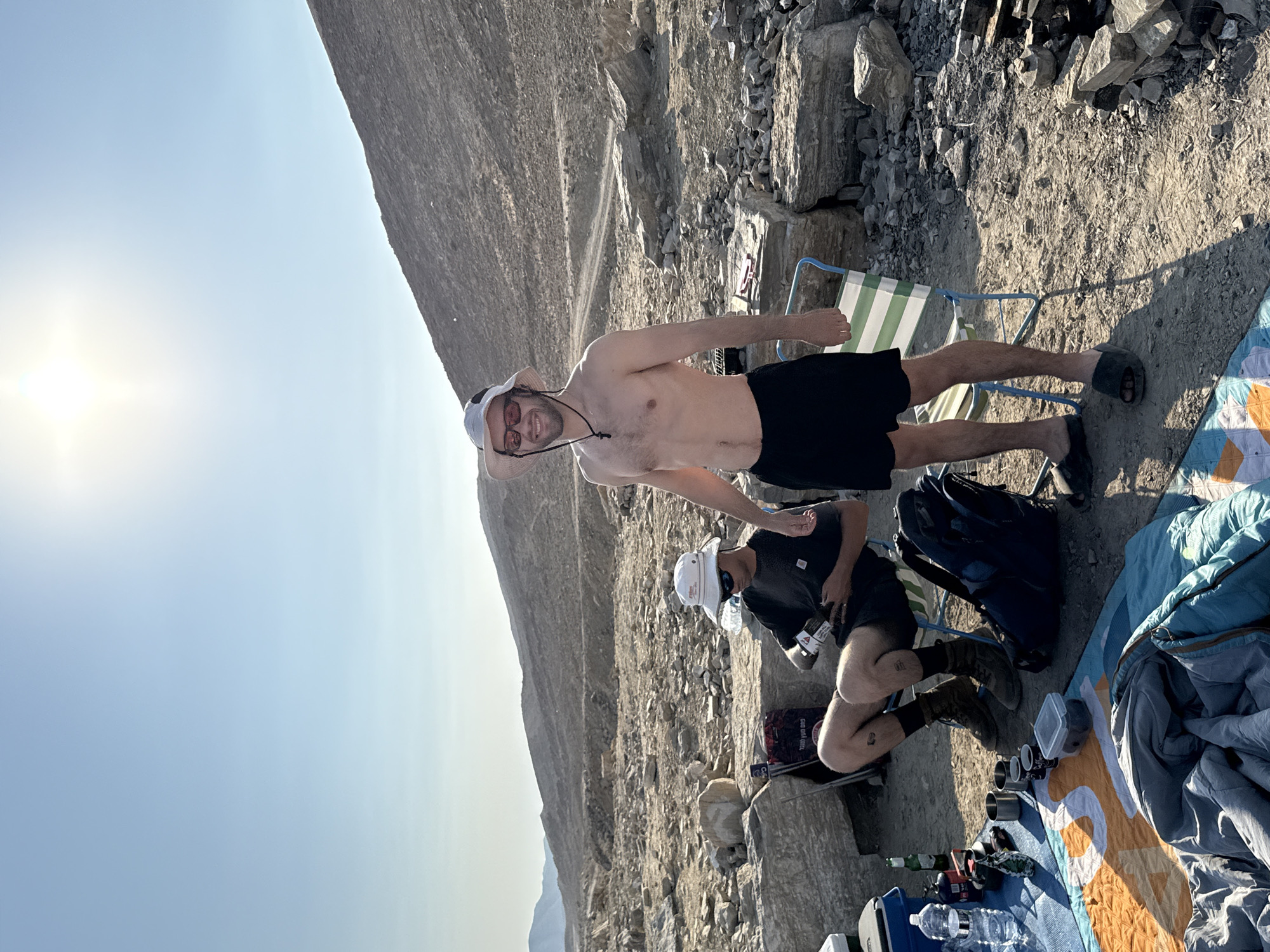 Two shirtless men, wearing bucket hats and sunglasses, are at a campsite in a rocky, desert terrain. The man on the right is standing and smiling, wearing black shorts. The man on the left is seated in a camping chair, wearing black shorts and boots, and holding a small object. The campsite includes camping chairs, a blanket, a sleeping bag, cups, bottles, and a backpack. In the background, a dusty hillside rises under a clear blue sky with the sun shining brightly. The location is identified as Ramat Negev Regional Council, Israel, characterized by arid, mountainous landscapes.