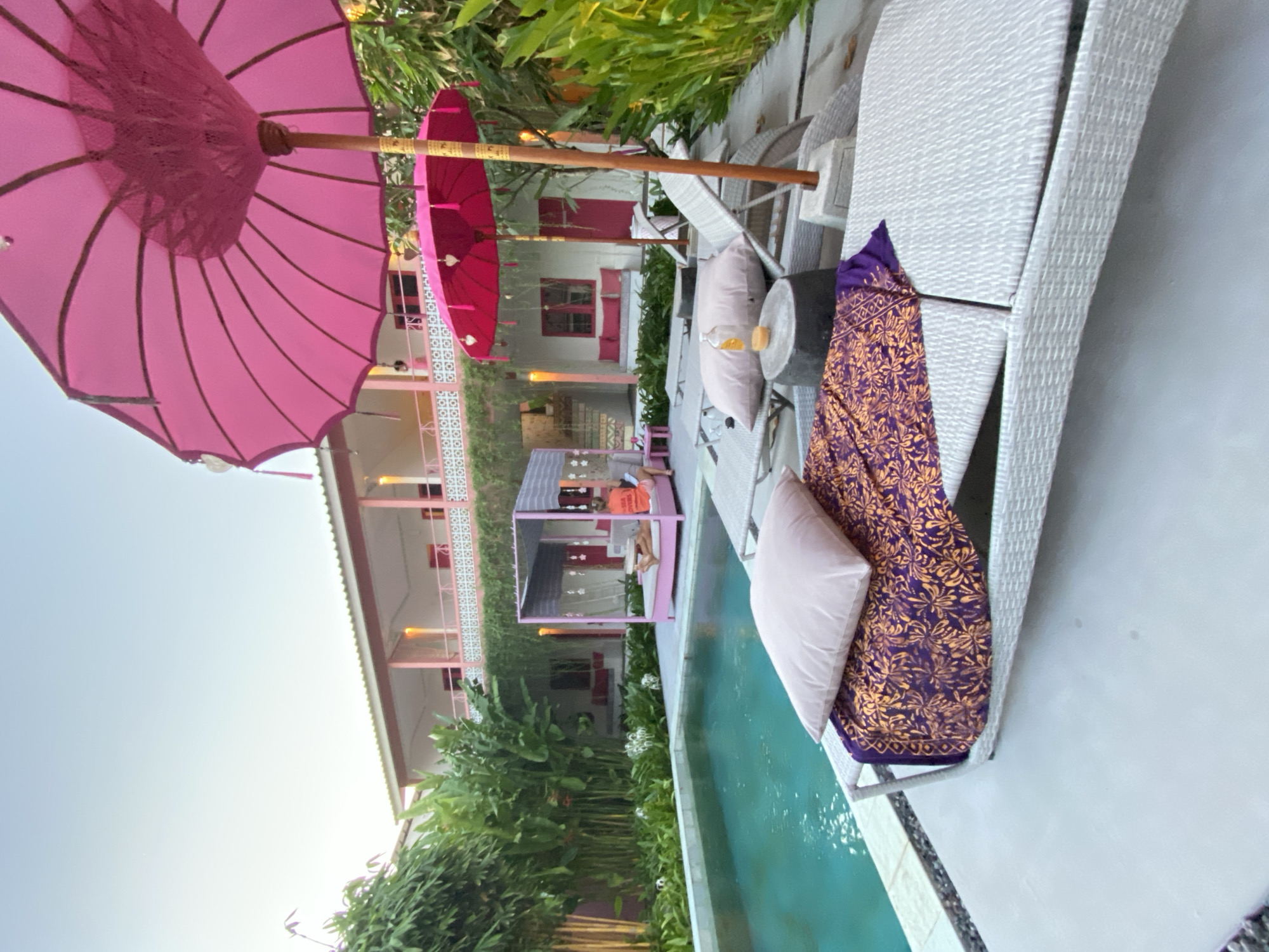 A low-angle, outdoor, wide shot captures a poolside scene with a building and lush greenery in the background. The foreground features a white, woven chaise lounge with a purple and gold patterned textile and a light pink pillow. To the left, a turquoise swimming pool is visible, bordered by grey stone. Behind the pool, a pink gazebo with sheer curtains encloses a pink daybed where a person is lying down. The building behind the gazebo has a white exterior with pink accents and balconies. Two large pink umbrellas are prominently displayed in the middle ground, casting shadows. The scene is illuminated by natural light.