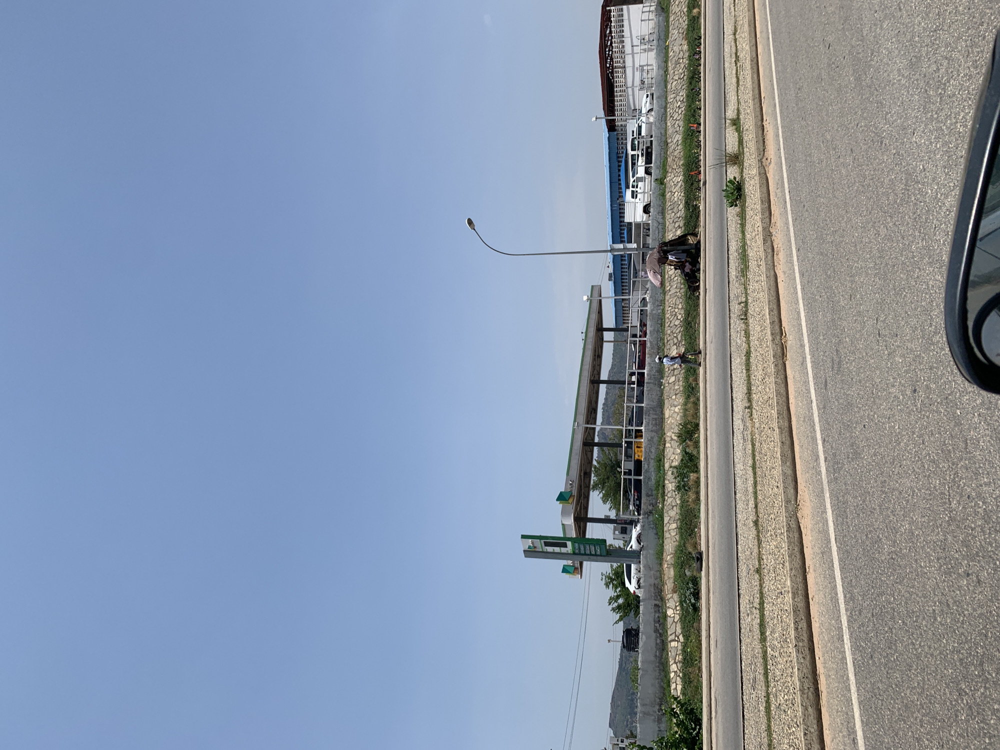 The image shows a roadside view in Kubwa, Nigeria. A gas station with a price sign and canopy is visible on the left side of the frame. The gas station is situated beside a building that houses multiple white vans. A street light stands nearby. Two people are standing near the building, one holding an umbrella. A grey road stretches along the right side of the image, with a car's side mirror visible in the foreground. The sky is a clear, pale blue.