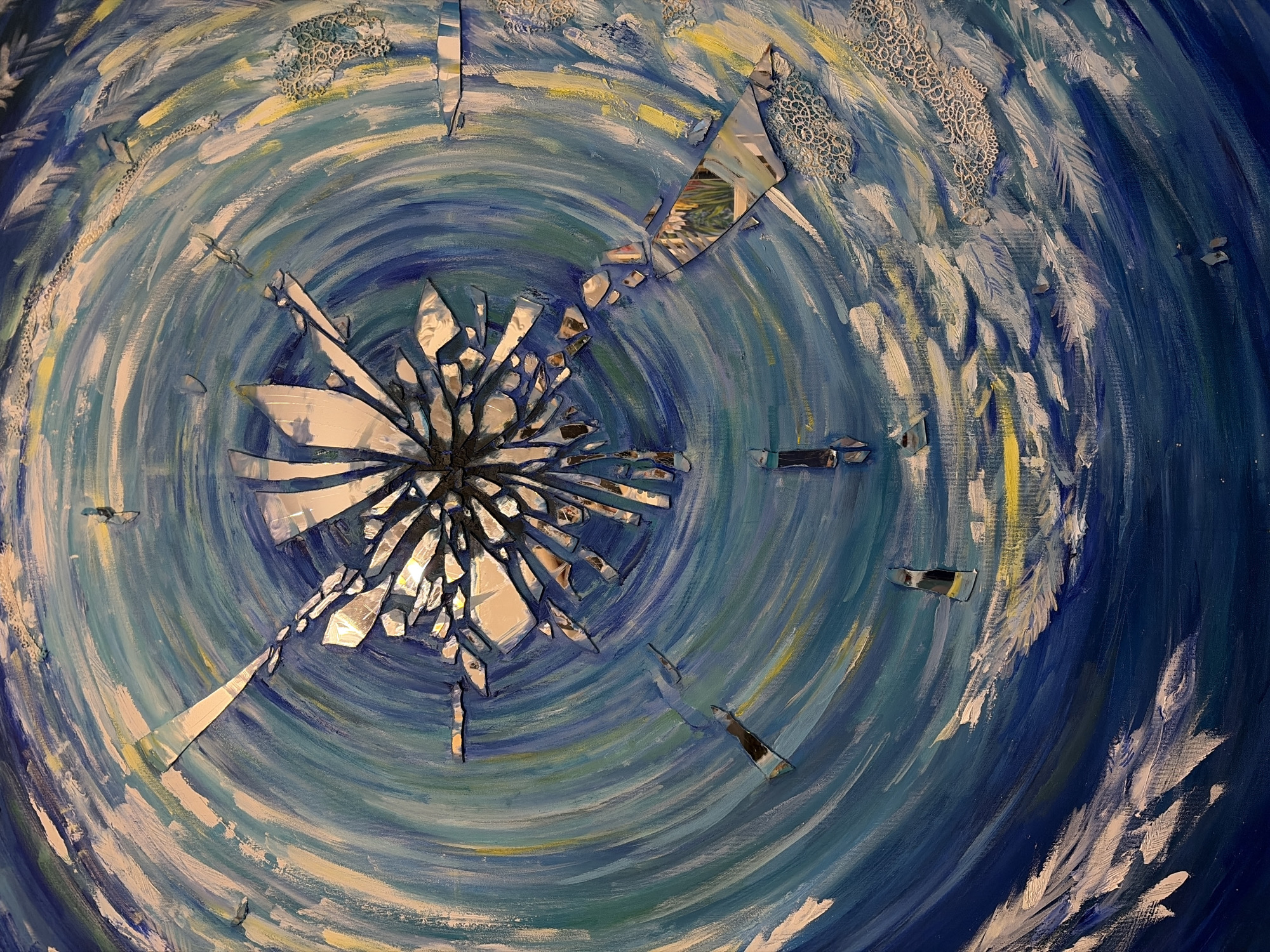 The image is a detail shot of an artwork, likely a painting or mixed-media piece, featuring a swirling, circular pattern dominated by blue and white hues, reminiscent of water or a vortex. At the center of the swirl is a fragmented, starburst-like arrangement of reflective shards, resembling broken mirrors. Some of these shards reflect external images, hinting at a real-world setting. The painting also incorporates textured elements, such as what appears to be lace or other fabric patterns, integrated into the painted surface. There is no way to verify from the images provided that the location is in Budva, Montenegro.