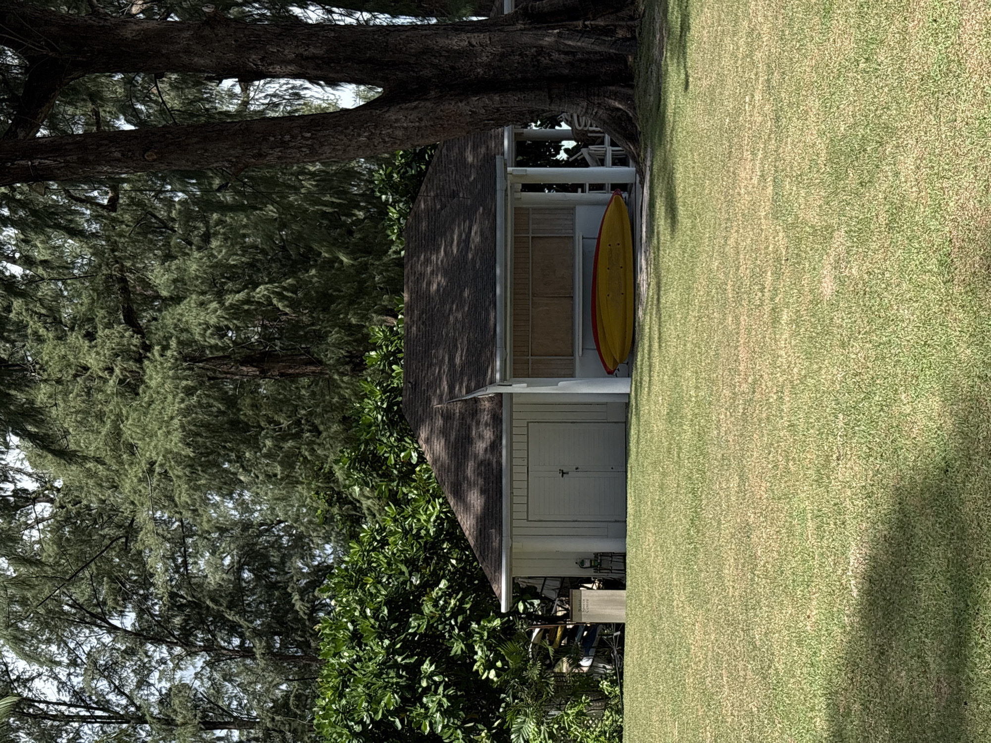 A modest, single-story building with a dark, sloped roof stands on a well-maintained lawn, surrounded by lush green foliage and tall trees. The building features white siding, a double door garage entrance on the left, and an open-air covered area on the right with two kayaks, one red and one yellow, resting on the ground. A sign that reads "Water Sports Centre" is visible next to some stacked watercraft on the left side of the building. The scene is bathed in natural daylight, casting shadows across the grass. The environment appears to be a serene outdoor recreational area. This scene is located in the city of Choeng Thale, Thailand.