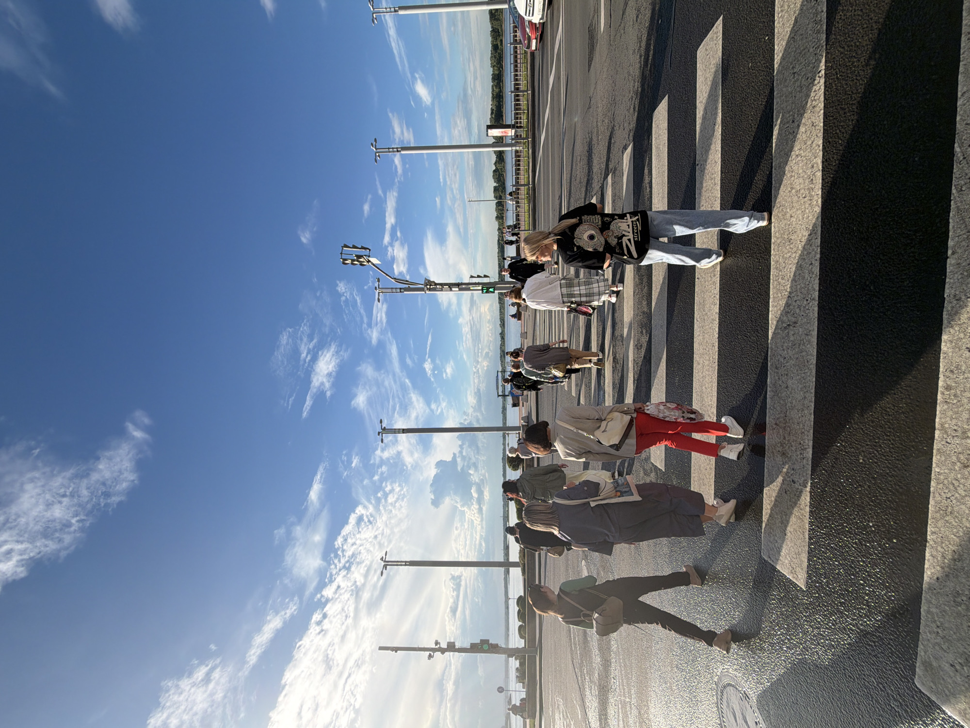 In Nizhny Novgorod, Russia, a group of people are crossing a pedestrian street under a clear blue sky with scattered white clouds. The sun is high, casting long shadows across the striped crosswalk, suggesting it is late afternoon or early evening. The scene depicts a street lined with traffic lights and poles, indicating an urban environment.
The individuals, seen from the waist down, are dressed in casual attire. Some are wearing jeans, t-shirts, and sneakers, while others are in skirts and trousers. One person is carrying a dark bag, and another has a light-colored purse. The people are walking in the same direction, suggesting they are all moving across the street. The mood appears neutral, with no strong emotional cues visible.
In the background, a body of water is visible, possibly a river, with trees and buildings on the far bank. This suggests the crosswalk is located near a waterfront area. A few cars are parked on the side of the road, and other people can be seen in the distance. The image captures a slice of everyday life in this Russian city, highlighting a moment of transit and the serene atmosphere of a sunny day.