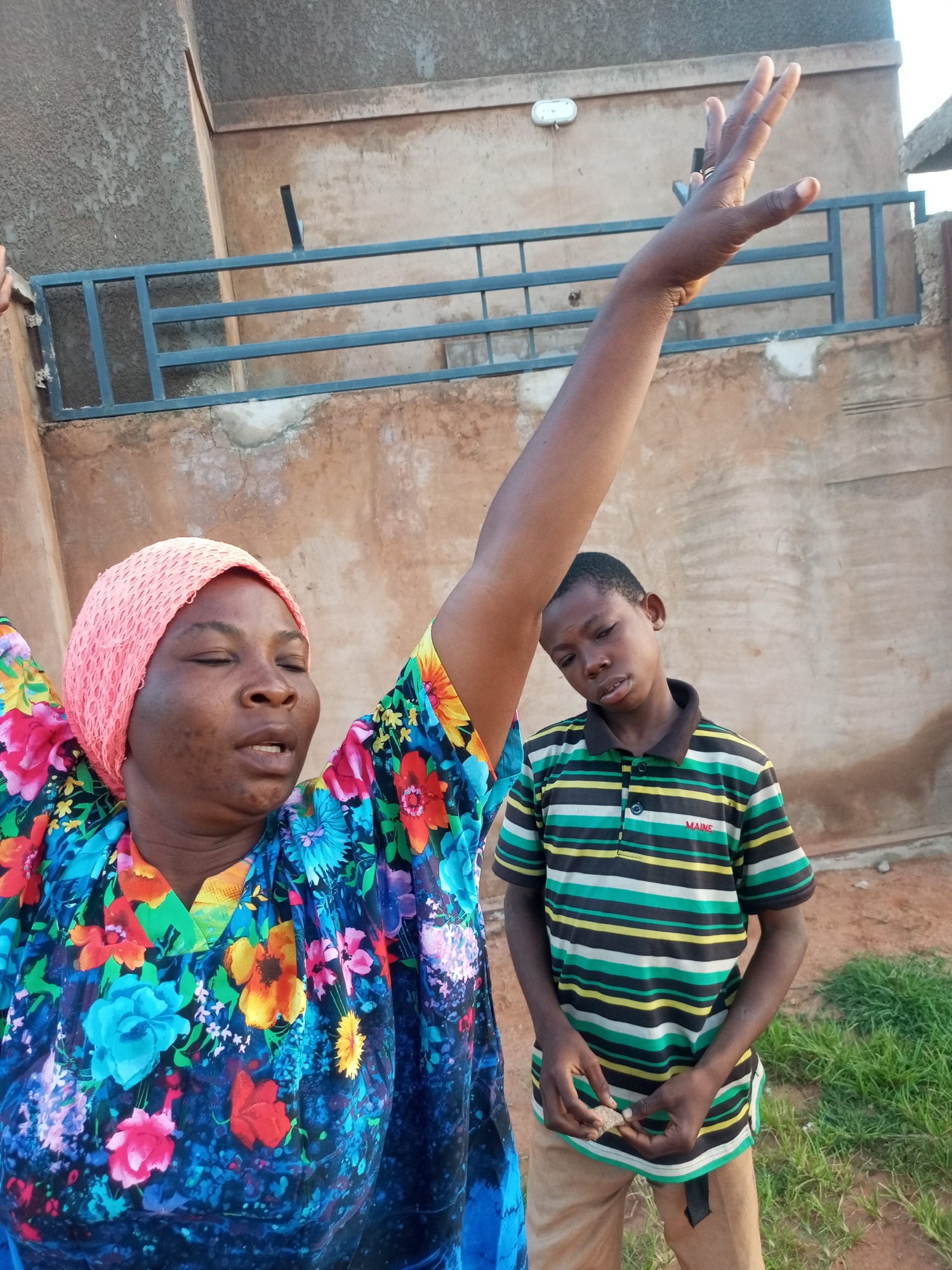 A medium shot captures a woman with her eyes closed and right arm raised in the air, seemingly in prayer or exaltation, standing in front of a young boy who stands with his head tilted and eyes downcast. The woman wears a bright floral-print dress and a pink headscarf. The boy is dressed in a striped t-shirt with "MAINE" written across it and tan pants. They are outdoors, with a textured, light brown building and a blue metal railing visible in the background. The ground appears to be dirt with patches of green grass. The scene is located in Tempure, Nigeria.