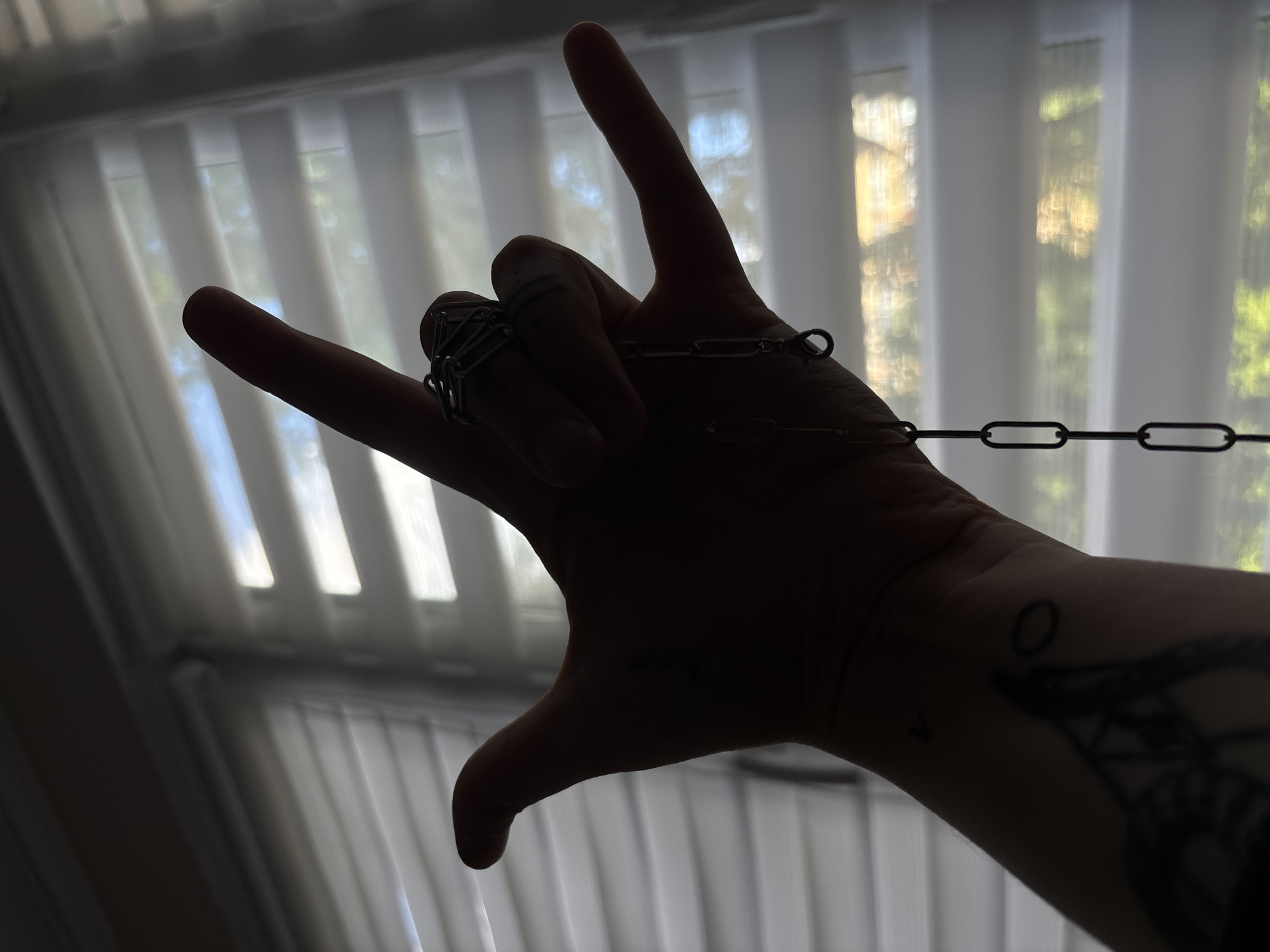 A silhouetted left hand is held up, fingers extended in a gesture that resembles the "rock on" symbol, with the thumb and index finger forming an "L" shape. A silver chain is draped from the ring finger, which is adorned with a large, multi-banded silver ring. The hand is positioned in front of a window with horizontal blinds, through which sunlight streams, creating a hazy, backlit effect. The overall environment is indistinct due to the backlighting. No specific details about Saint Petersburg, Russia, are discernible in the image.