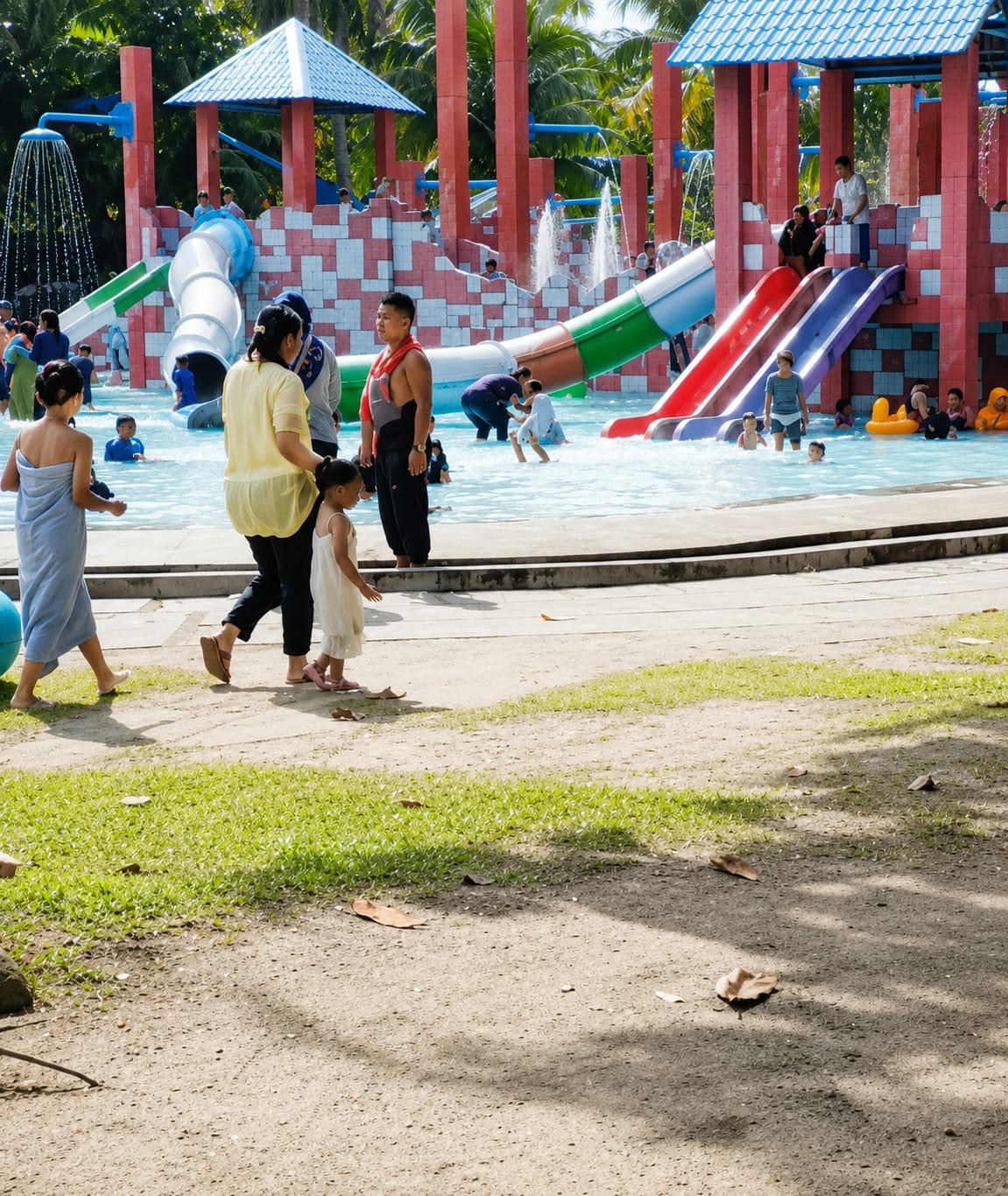 A group of people, including adults and children, are enjoying a sunny day at a water park. In the foreground, a woman in a yellow top and black pants walks with a young girl in a white dress. To their left, another woman in a blue towel walks away from the camera. In the background, there is a large, colorful water slide structure with multiple slides, a pool, and fountains. People are seen playing in the water and on the slides. The environment appears to be a tropical setting with palm trees visible. The location is specified as Pantai Cermin Kiri, Indonesia.