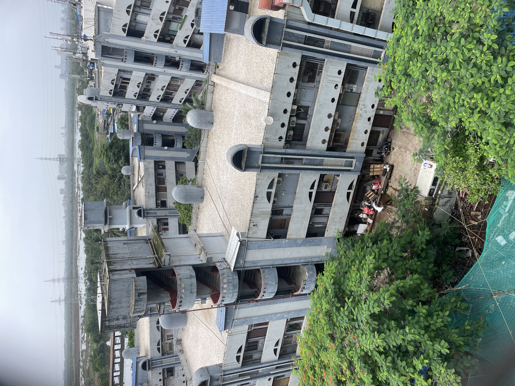 This is an aerial, eye-level medium shot of a cluster of aged, Brutalist-style apartment buildings in the Mumbai, India metropolitan area. The buildings, which appear to be constructed of concrete and painted a light gray, exhibit signs of wear and tear, including discoloration and some moss growth. The architecture is characterized by strong geometric shapes, with protruding sections and rounded elements. An exterior spiral staircase with red accents winds up the side of one of the buildings. The rooftops are flat and covered in a light-colored aggregate material, with various pipes, vents, and satellite dishes present. In the foreground and surrounding the buildings are lush green trees, some with small red flowers. Beyond the buildings, a body of water is visible, with more vegetation and distant city skyline, including power lines and tall buildings, under an overcast sky. At the base of the buildings, a small courtyard area shows some debris, wooden pallets, a white van, and a scooter with two people. A green safety net is draped over some construction materials in the lower left corner.
