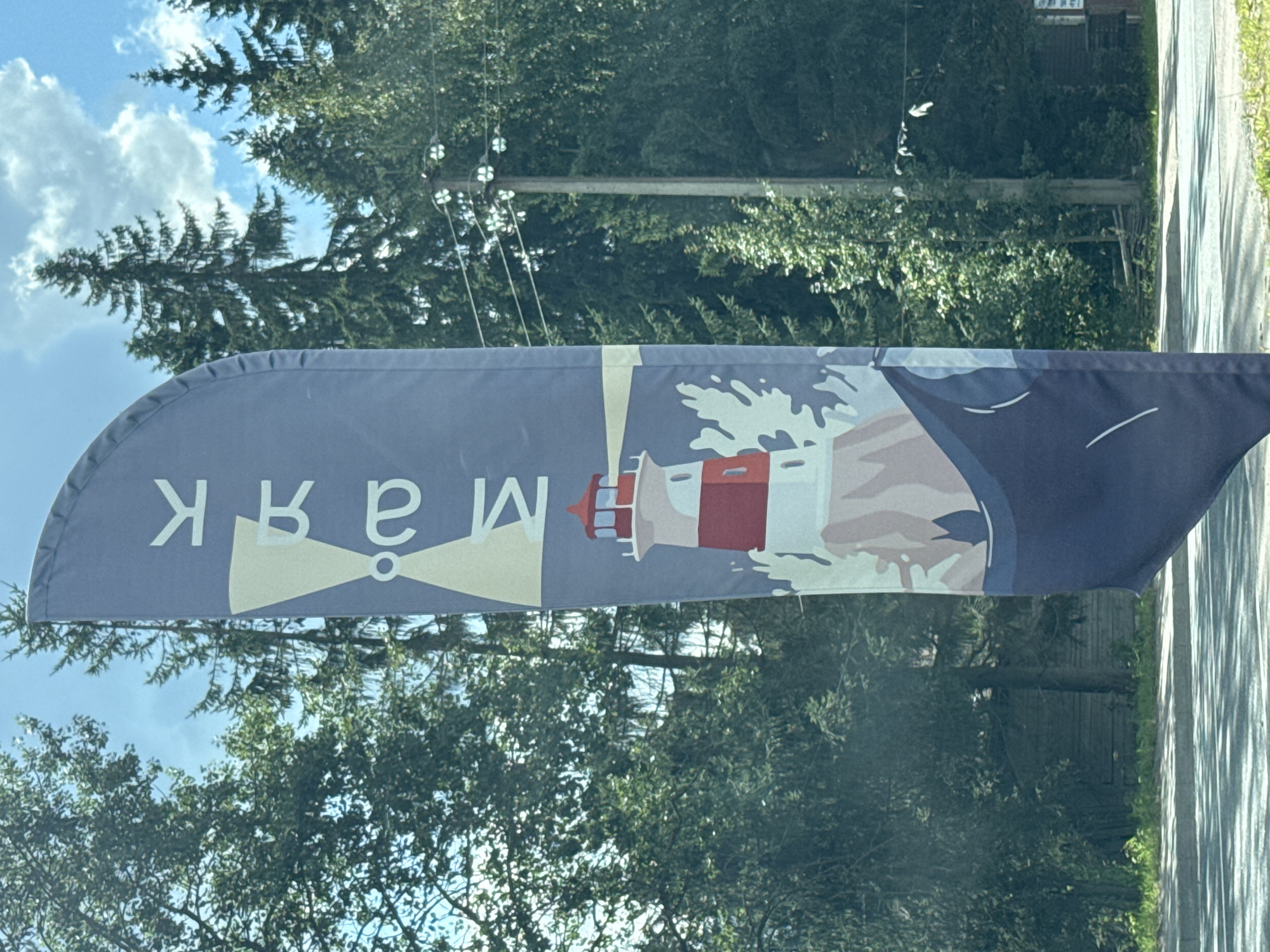 A vertical banner displaying a lighthouse graphic and text in Cyrillic is positioned in front of a backdrop of trees and a cloudy sky. The banner is predominantly blue with white and light yellow accents. The lighthouse is red and white and is depicted on a rocky outcrop with waves. Above the lighthouse, there are two white shapes that resemble propellers or wings, with a white circle in the center. The Cyrillic text reads "МАЯК", which translates to "Lighthouse". The banner is fluttering, indicating a breeze. In the background, tall evergreen trees form a dense forest. The sky is blue with scattered white clouds. The foreground shows a paved surface, possibly a road or sidewalk, with some greenery on the left side. The scene appears to be outdoors in a natural setting, likely in or near the city of Yakkima, Russia, given the presence of the Cyrillic text and the type of trees commonly found in such regions. There are no people or specific actions visible in the image.