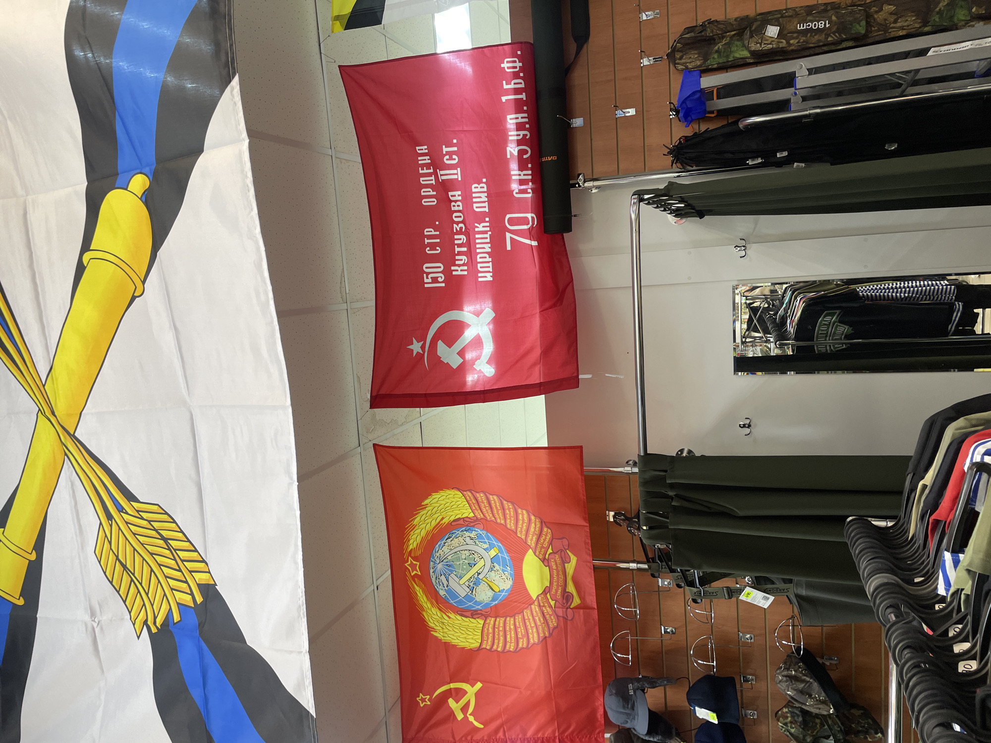 This media file captures an indoor view of a retail store in Krasnoyarsk, Russia, specializing in military-themed or outdoor gear. No people are visible.
Three large flags hang from the ceiling. On the far left is a white flag featuring a black and blue striped background with a golden cannon crossed with two golden arrows or quivers. To its right, closer to the center, is a red flag displaying a white hammer and sickle and a star in the bottom left, with extensive Cyrillic text in the center and top right. This text reads: "150 СТР. ОРДЕНА КУТУЗОВА II СТ. ИДРИЦК. ДИВ. 70 СТ.К. З.У.А.1Б.Ф." (150th Rifle Order of Kutuzov 2nd Class Idritskaya Division 70th Str.K. Z.U.A.1B.F.), identifying it as a replica of the Victory Banner. Below this, another red flag hangs, featuring the emblem of the Soviet Union: a globe with a hammer and sickle, surrounded by a wreath of wheat, a rising sun, and the motto "ПРОЛЕТАРИИ ВСЕХ СТРАН СОЕДИНЯЙТЕСЬ!" (Proletarians of all countries, unite!) repeated in various languages, along with a smaller hammer and sickle and star in the bottom left.
To the right, the store features wooden slat walls with numerous hooks displaying various items. Metal clothing racks hold an assortment of apparel, including olive green trousers, black shirts, and other garments. A camouflage bag marked "180cm" and a black case with "LVO" are visible on the upper shelves. Below, several hats are displayed, some with tags, one bearing the brand "SARDONIX." A mirror on the back wall reflects more clothing. The setting suggests a military surplus or outdoor goods store.