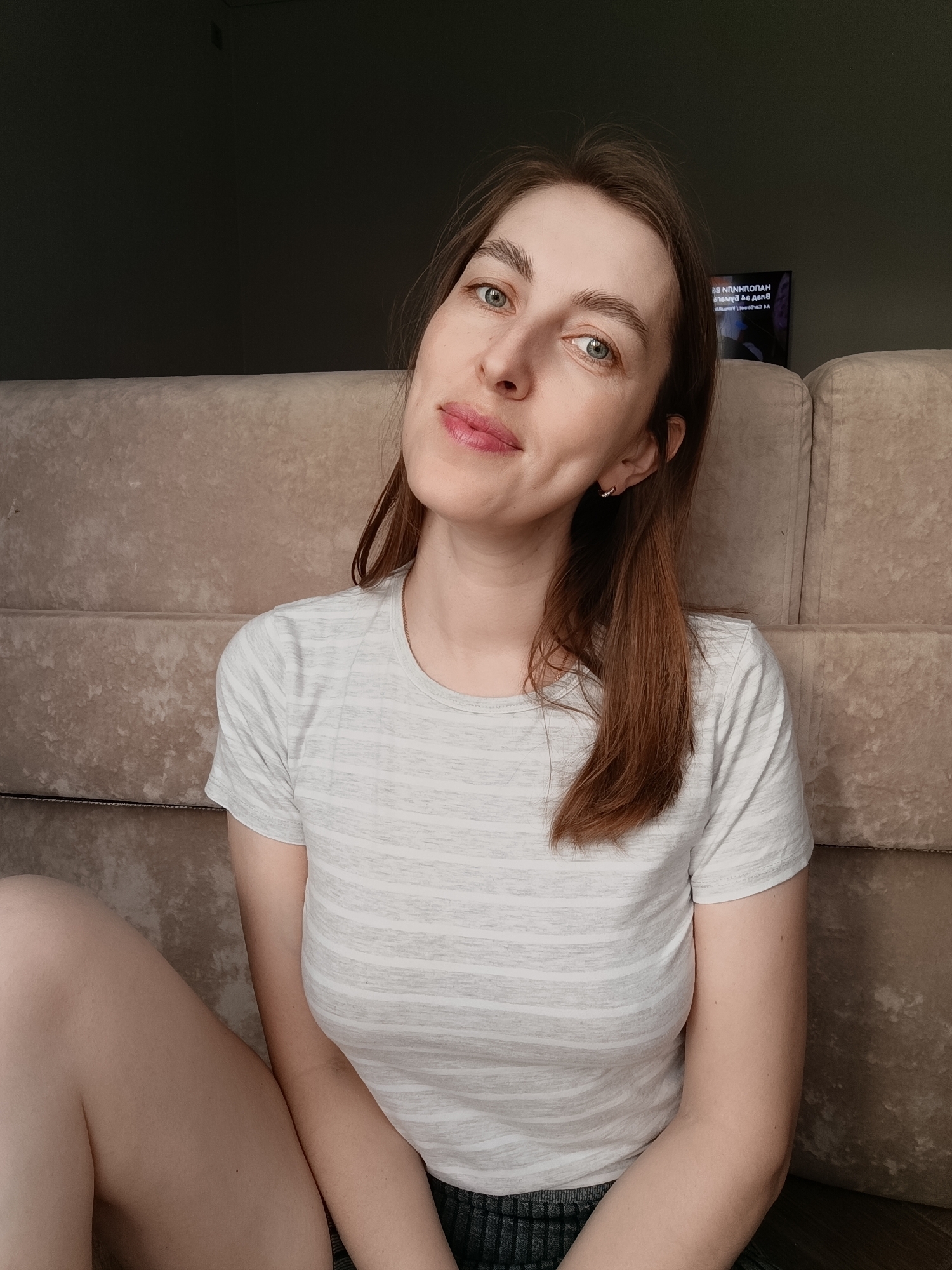 A woman with light skin, green eyes, and light brown hair is sitting on a beige couch. She is wearing a light gray striped t-shirt. Part of her bare left leg is visible. The background is a dark wall, and a television is visible in the upper right corner of the frame. The location is Chaikovsky, Russia.