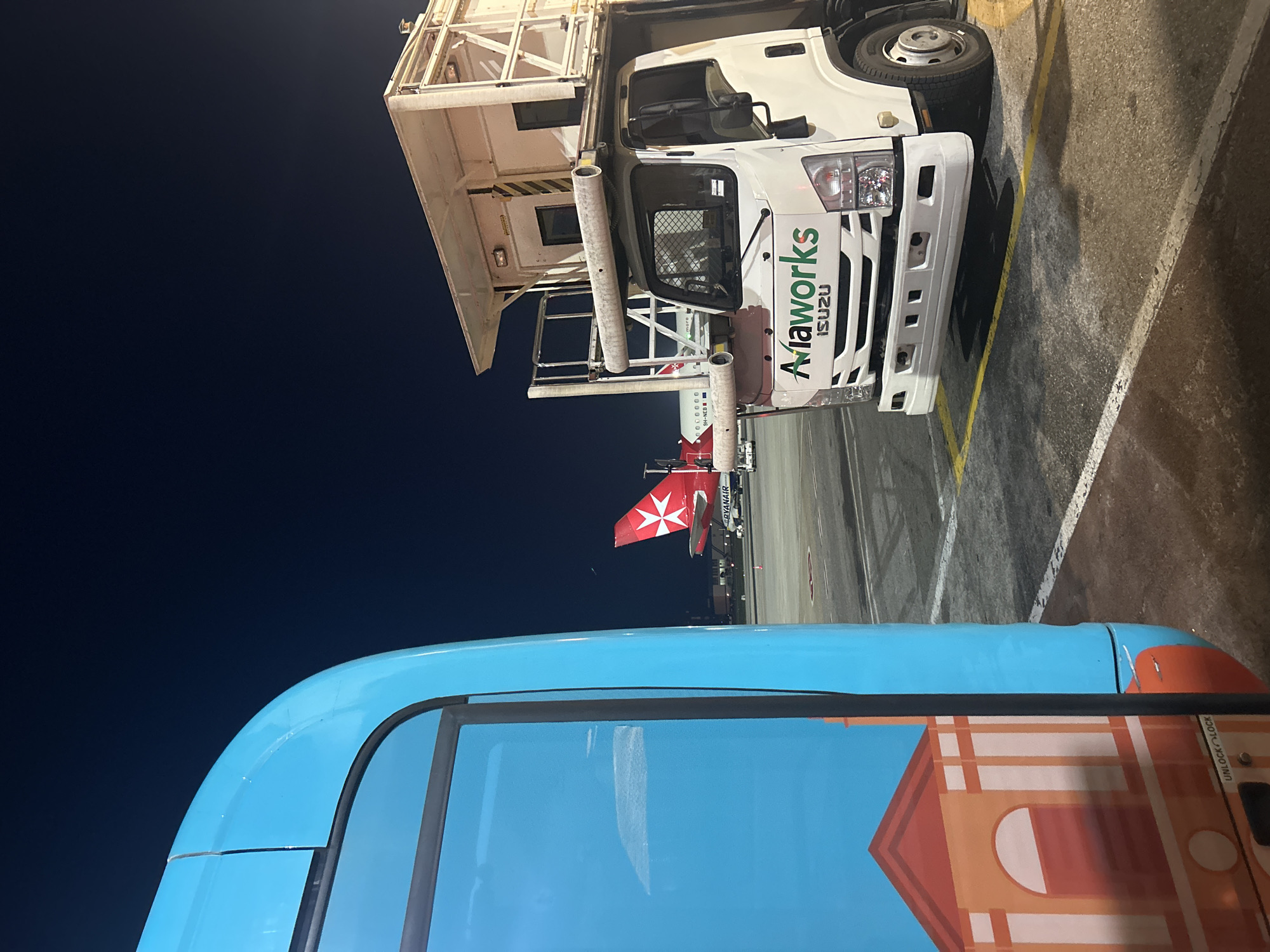 The image depicts an airport scene in Luqa, Malta, at night. A white Isuzu "Aiaworks" catering truck with a platform is parked on the tarmac. In the background, a Ryanair aircraft with the Maltese eight-pointed cross on its tail fin is visible. The foreground features a blue bus with a graphic of a building. The ground is paved with asphalt, and there are painted yellow lines indicating parking. The sky is dark.