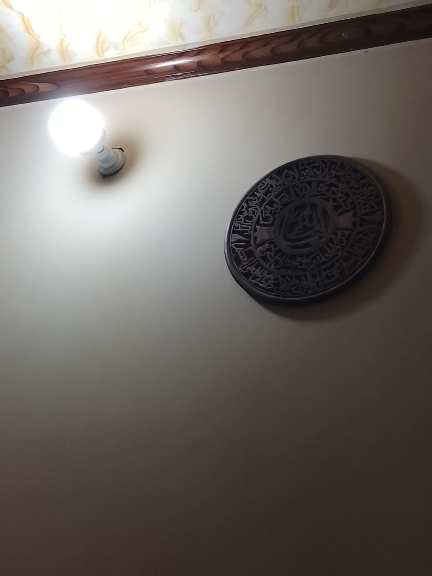 A brightly lit interior wall is visible, with a light fixture in the upper left and a circular wall hanging to the right. The wall is a light beige color. Above the wall is a dark brown trim. The wall hanging is circular and appears to be made of a dark material with Arabic calligraphy inscribed on it. The photo was taken indoors in Multan, Pakistan.