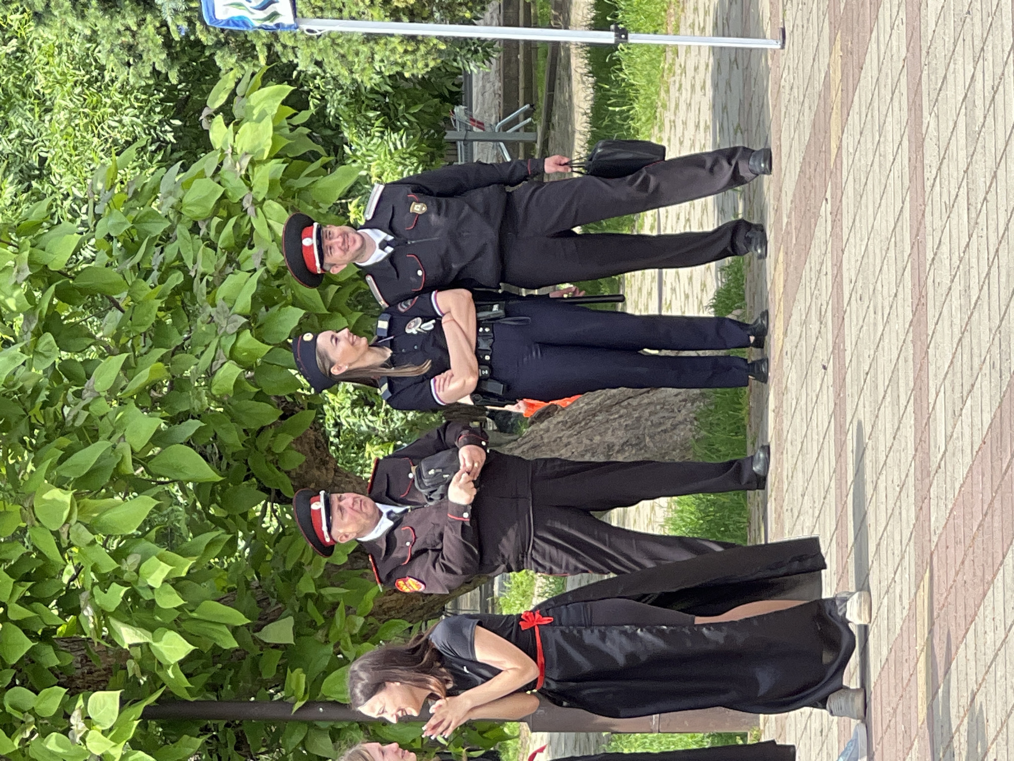 The image shows three individuals in formal law enforcement attire, and another individual in a long black dress with a red bow, posing outdoors on a paved area. The scene appears to be located near a tree, possibly in a public space in Goryachiy Klyuch, Russia. The people are standing facing the camera, two men in traditional Russian Cossack uniforms and one woman wearing a police uniform. All are wearing hats. The woman and the woman in the black dress are looking at one another, while the men are looking straight ahead.