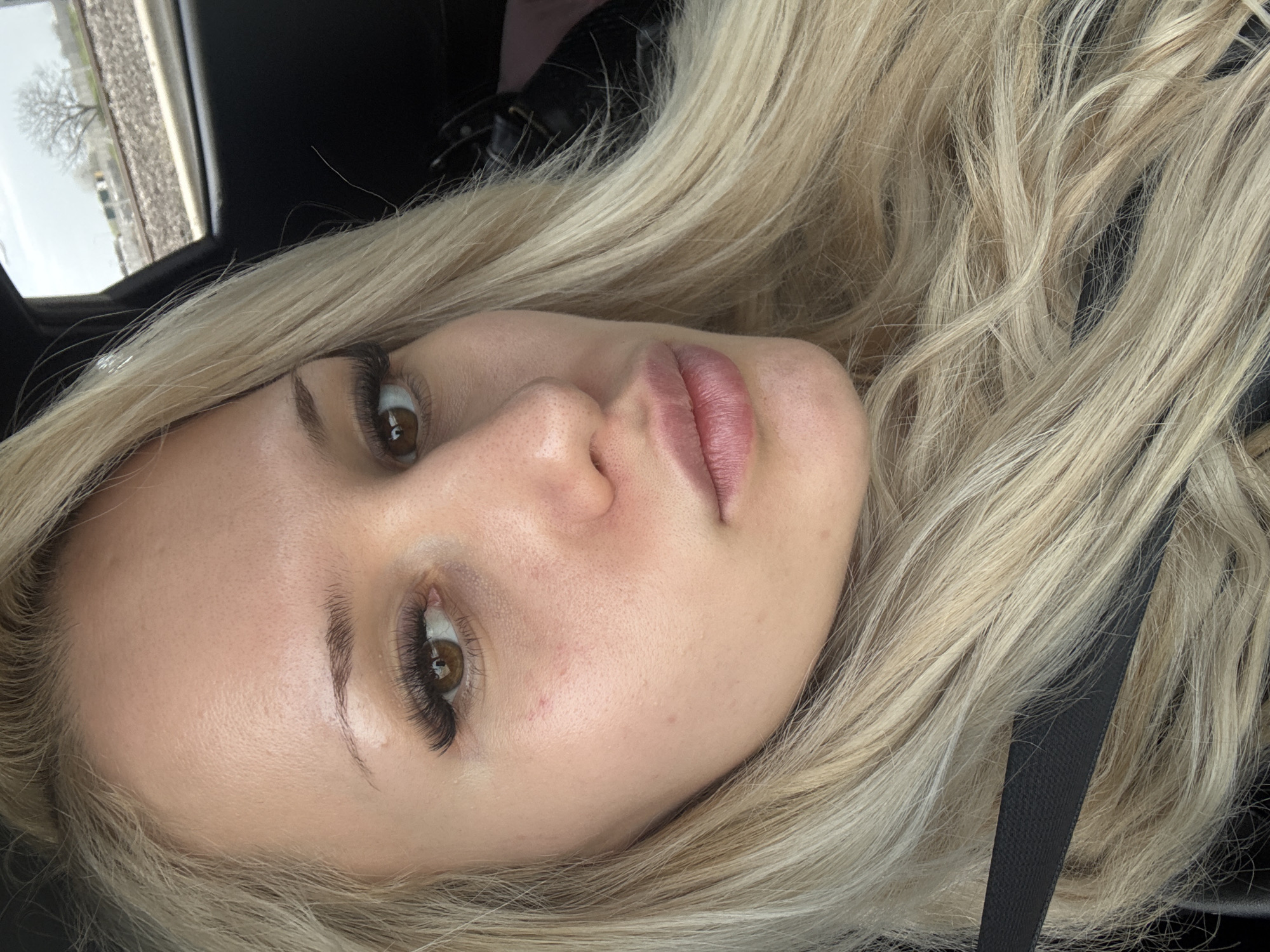 The image presents a close-up, head-and-shoulders portrait of a fair-skinned female individual with long, wavy blonde hair. Her brown eyes are open, featuring enhanced eyelashes, and she looks directly at the camera. Her lips are full and slightly parted. A dark, textured seatbelt strap diagonally traverses the lower left quadrant of the frame, partially obscured by her hair. The individual is situated within a dark interior, consistent with a vehicle. Through a window in the upper right background, railway tracks with ballast are visible under an overcast sky. A bare deciduous tree stands in the middle distance beyond the tracks, with indistinct industrial or urban structures visible in the far background. The scene is located in the city of Skutare, Bulgaria.