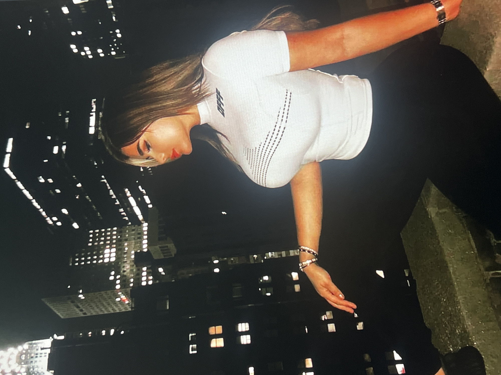 A young woman with long, dark hair and blonde highlights, wearing a white, short-sleeved, mock-neck top with a graphic print and black leggings, is seated on a concrete ledge against a nighttime cityscape. The background features illuminated skyscrapers with numerous lit windows, suggesting a dense urban environment. The woman is looking to her right, with her head tilted slightly. She has on bold red lipstick and dark eyeliner. Her right arm is extended forward, resting on the ledge, adorned with bracelets. Her left arm is bent at the elbow, with her hand resting on her lap. The overall lighting is dim, with the city lights providing the primary illumination. The image appears to be a photograph taken at night.