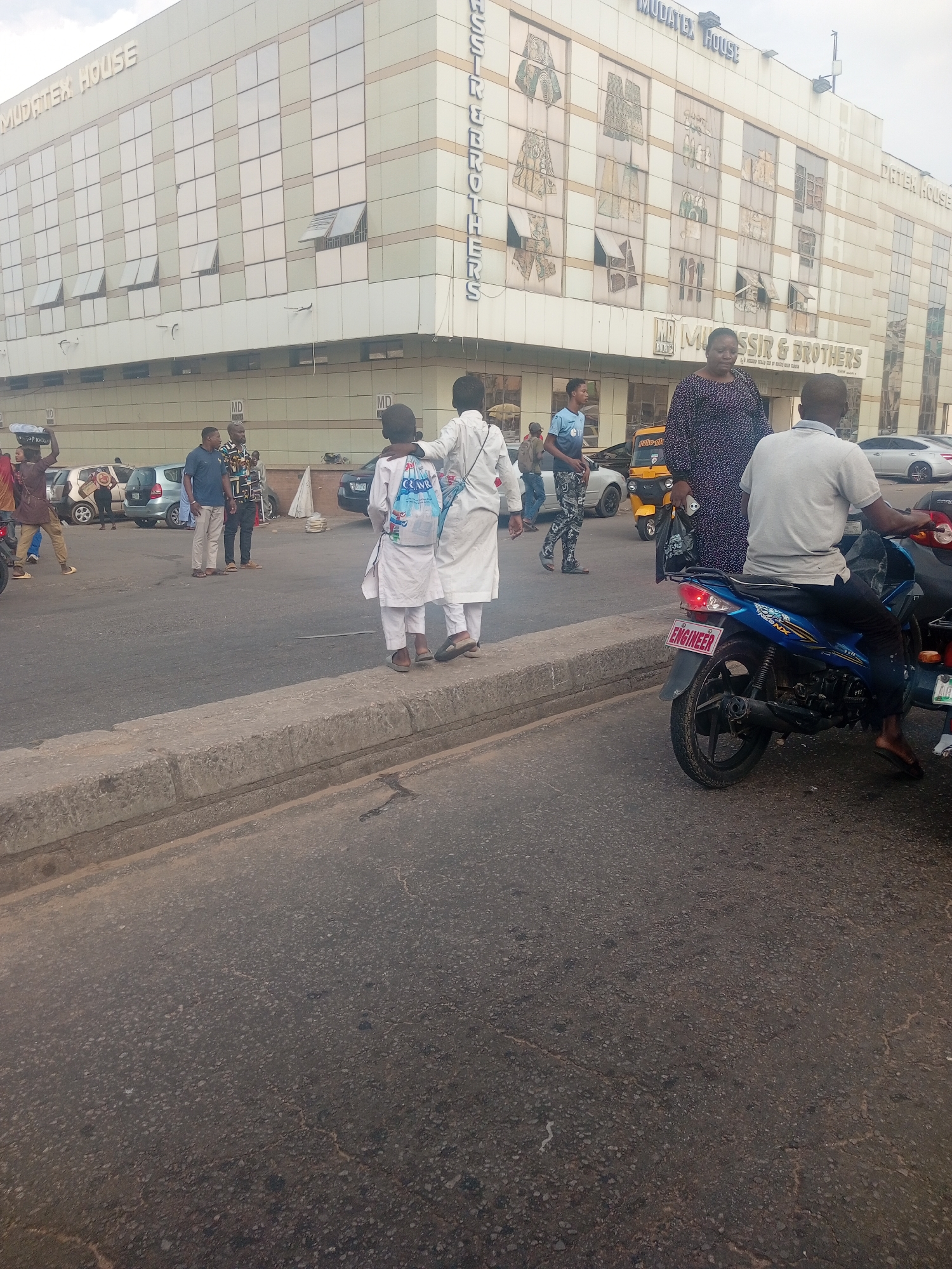 In Maiburiji, Nigeria, a street scene unfolds in front of the Mudatex House, a modern, multi-story building with a facade of reflective glass panels. The building features prominent signage that reads "MUDATEX HOUSE" and "MD MUSSIR & BROTHERS." The street is paved with asphalt and is lined with a concrete curb. Several people are present: two young boys in white garments stand near the curb, one with a backpack, facing away from the camera; an adult male in a gray polo shirt and dark pants is seated on a blue motorcycle, with a license plate displaying "ENGINEER"; to the left, a group of people are walking and standing. Among them, a man in a blue shirt and light-colored pants stands with another man in a patterned shirt. Further left, a woman carries an object on her head, and other individuals are visible near parked cars, including a yellow auto-rickshaw. The overall environment appears to be an urban commercial area.