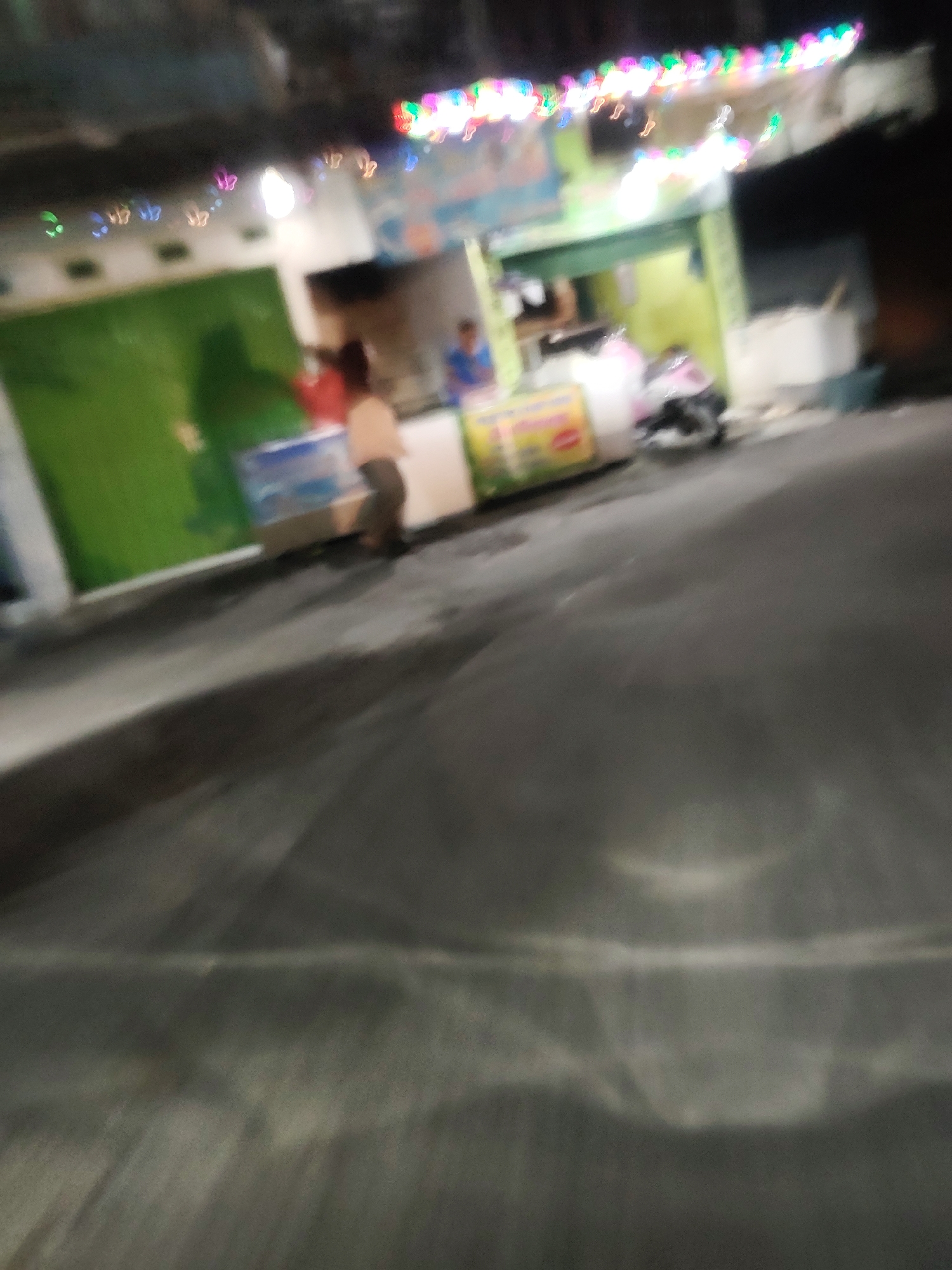 The image depicts a blurry nighttime scene outside a food stall in Mekarmukti, Indonesia. The stall has a green facade and is illuminated by colorful string lights across the top. A person, possibly a vendor, stands behind a counter, and another individual is partially visible to the left. A motorcycle is parked in front of the stall. The foreground shows a dark, wet asphalt road with reflections from the lights. The overall impression is one of a typical street-side eatery at night, rendered indistinct by motion blur.