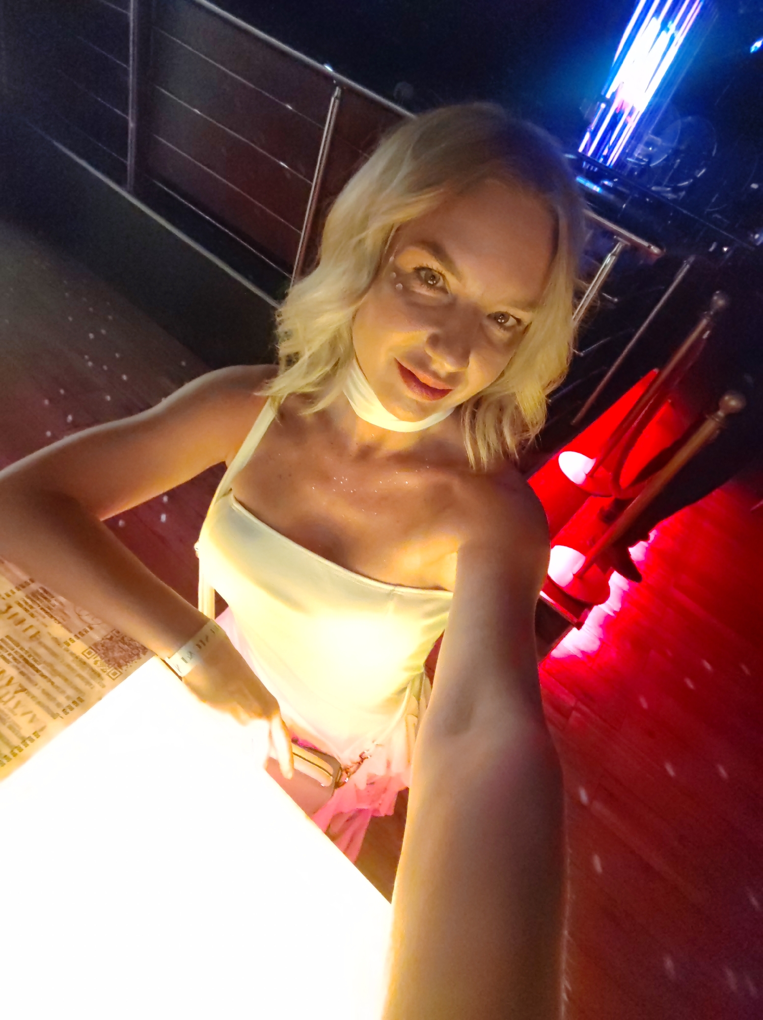 The image features a woman with blonde hair, looking at the camera with a slight smile. She is wearing a light-colored, strapless top and appears to have some glitter on her chest and collarbone. She has on a white wristband. The background is dark and features red and blue lights, suggesting a nightlife setting like a club or bar. The floor has a wooden texture, and there is a railing in the upper part of the image. To the left, there is a brightly lit surface, possibly a table, with some text and a QR code visible on what appears to be a menu. The overall ambiance is festive and energetic, consistent with a night out.