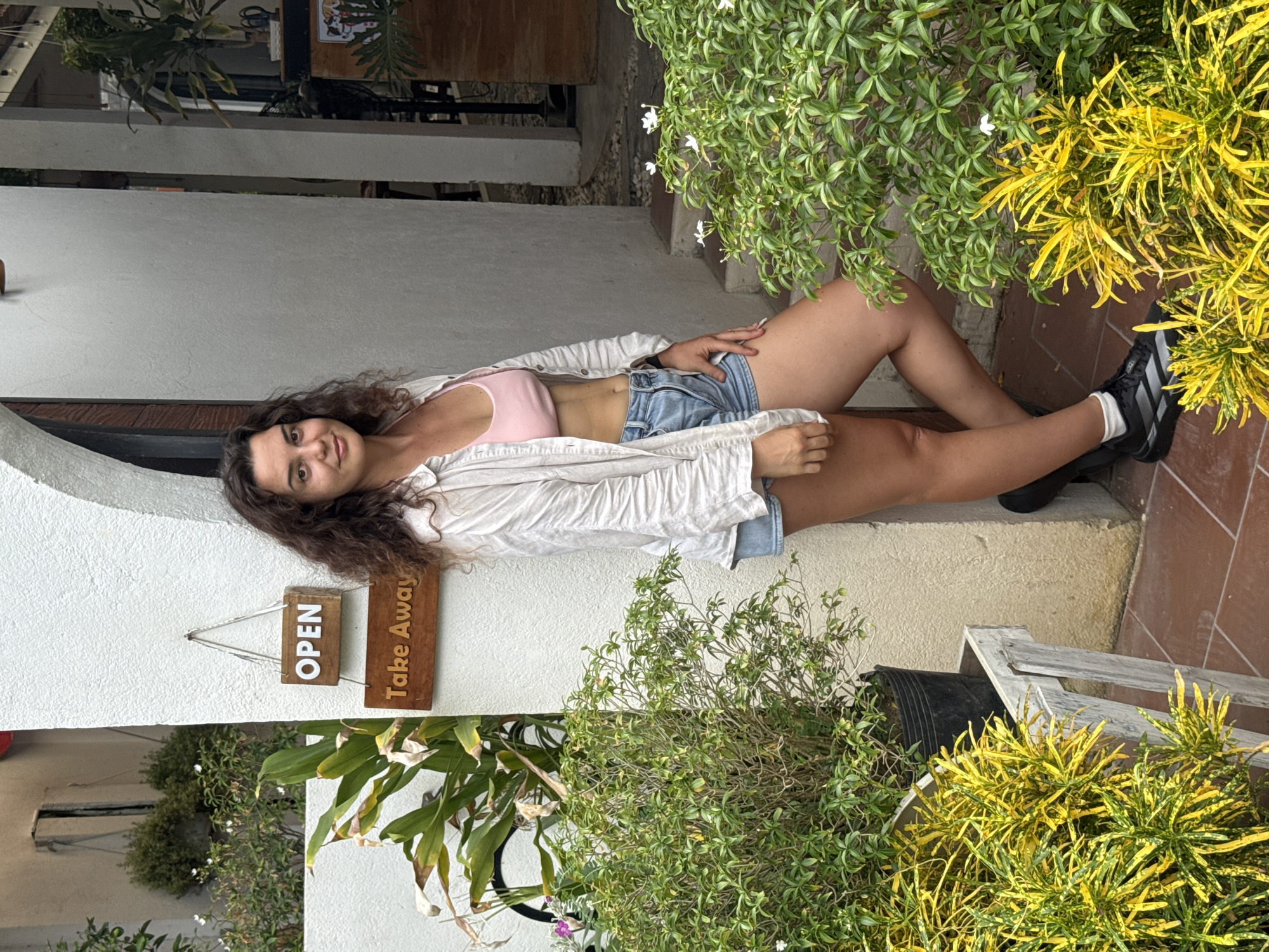 A young woman with dark, curly hair stands in front of a white wall with an arched opening. She is wearing a light-colored, open button-down shirt over a pink crop top and denim shorts. She has on black sneakers with white socks. Two wooden signs hang on the wall next to her. The upper sign reads "OPEN" in white lettering, and the lower sign reads "Take Away" in carved lettering. She is leaning against the wall and looking towards the camera. In front of her, there are various plants, including a potted plant with large green leaves and a smaller plant with bright yellow and green foliage. The ground is tiled with reddish-brown tiles. The background shows an outdoor area with more plants and architectural elements, suggesting a cafe or restaurant setting. The scene is consistent with the common aesthetic of establishments in Hoi An, Vietnam, which often feature outdoor seating, lush greenery, and traditional architectural elements.