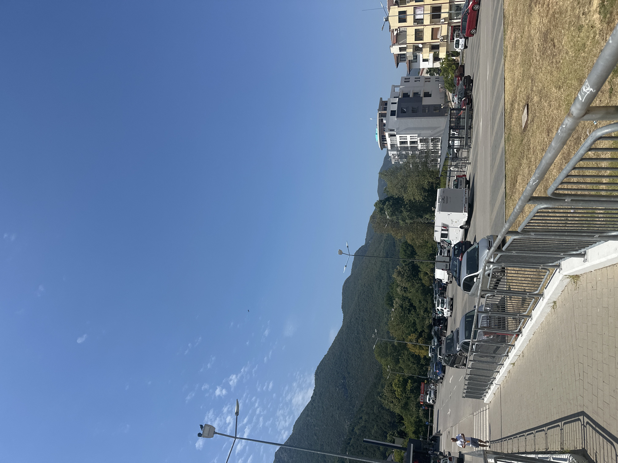The image depicts a sunny day in the city of Vratsa, Bulgaria, with a clear blue sky and scattered clouds. In the foreground, a paved pedestrian walkway runs alongside a metal fence. Beyond the fence, a busy street is visible with various vehicles, including cars, a camper van, and a white van. The street is lined with trees and buildings of different architectural styles. In the background, a large, green mountain dominates the landscape. Two pigeons are perched on a lamppost in the upper left corner of the image. A person is walking across a crosswalk on the left side of the image, carrying a white object. The scene appears to be a commercial or residential area within the city.
