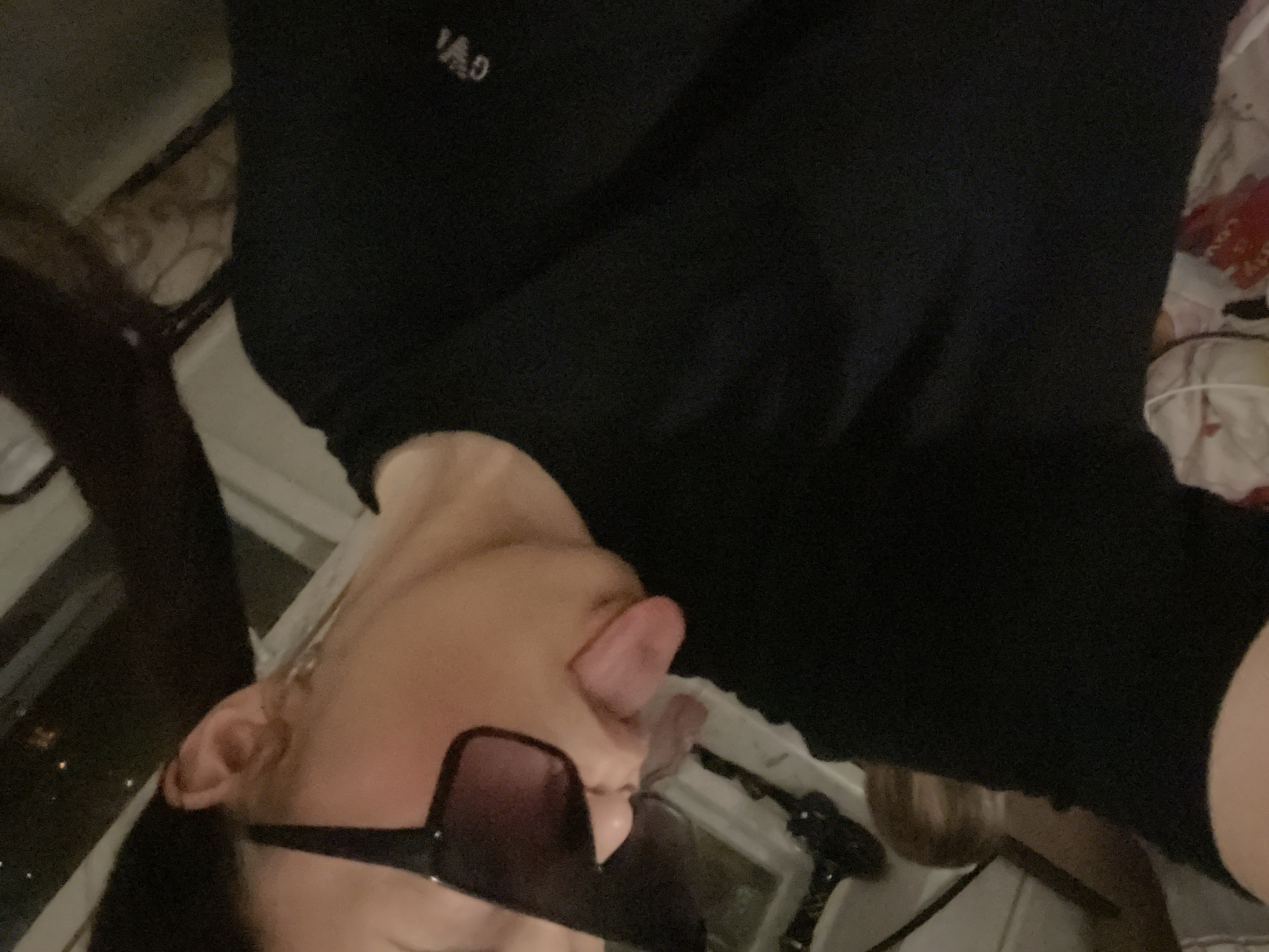 The image features a person, likely female, captured in what appears to be a selfie or close-up photo. Their head is tilted to the side, and they are wearing dark sunglasses that cover their eyes. A notable detail is their tongue playfully sticking out. The person is dressed in a plain black top or sweatshirt, upon which the faint, light-colored text "VAD" is visible on the chest area.
The scene suggests an indoor setting, possibly a room like a bedroom or living area. The lighting is dim, indicating it is likely night or late evening. Through a window or opening in the background, blurred lights that resemble distant city lights can be seen, hinting at an urban or suburban environment in Novoe Devyatkino, Russia. Portions of a patterned fabric, possibly bedding or a cushion, are also visible, along with other indistinct indoor elements. The overall impression is one of a casual, playful, or mischievous moment.