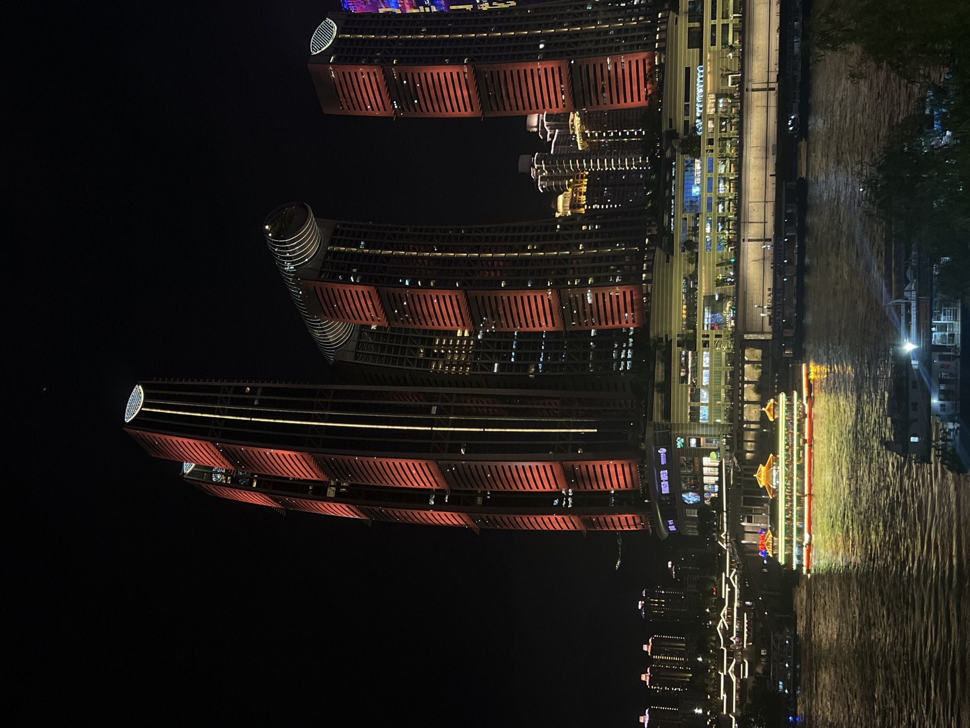 This is a night time photograph of a waterfront in Jiangbeicheng Subdistrict, China, featuring illuminated skyscrapers and a boat on the water. The prominent structures are several modern skyscrapers with their facades lit in red and yellow. The tallest building on the left has a distinctive rounded top with a circular pattern of lights. To its right, a curved skyscraper also displays red and yellow lighting. Further to the right, another skyscraper with red lighting is visible. In the foreground, a wide river flows, reflecting the lights from the buildings and a brightly lit traditional-style boat. The boat is adorned with green and yellow lights along its sides and has a distinct, multi-tiered roof structure. Several other smaller boats are present on the water. In the background, a cityscape of lower buildings with illuminated windows and outlines can be seen, suggesting a densely populated urban area. The overall scene is one of a vibrant and illuminated city at night.