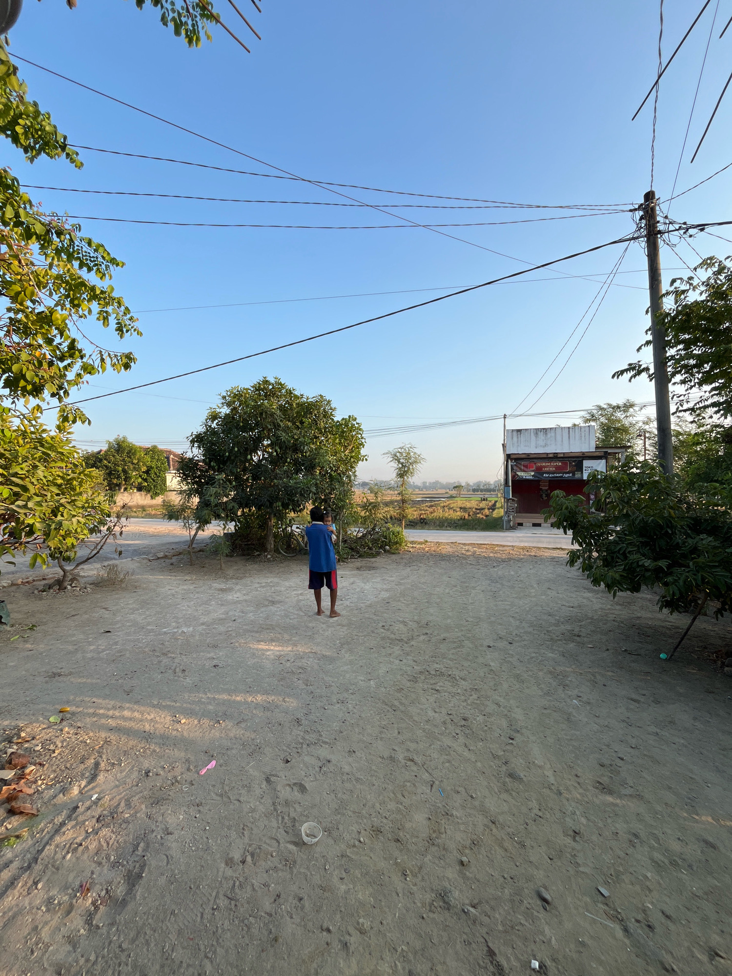 In Kauman, Indonesia, a young boy wearing a blue t-shirt and red shorts stands in a dirt courtyard, facing away from the camera and holding a child. The courtyard is strewn with debris including rocks, leaves, and a pink plastic object. Trees with green foliage are visible on either side of the frame. In the background, a building with a sign that reads "DURRUM SUPER KAEFEK" is visible, and beyond that, a field of crops under a clear blue sky. Overhead, power lines crisscross the sky, supported by a utility pole on the right side of the image.
