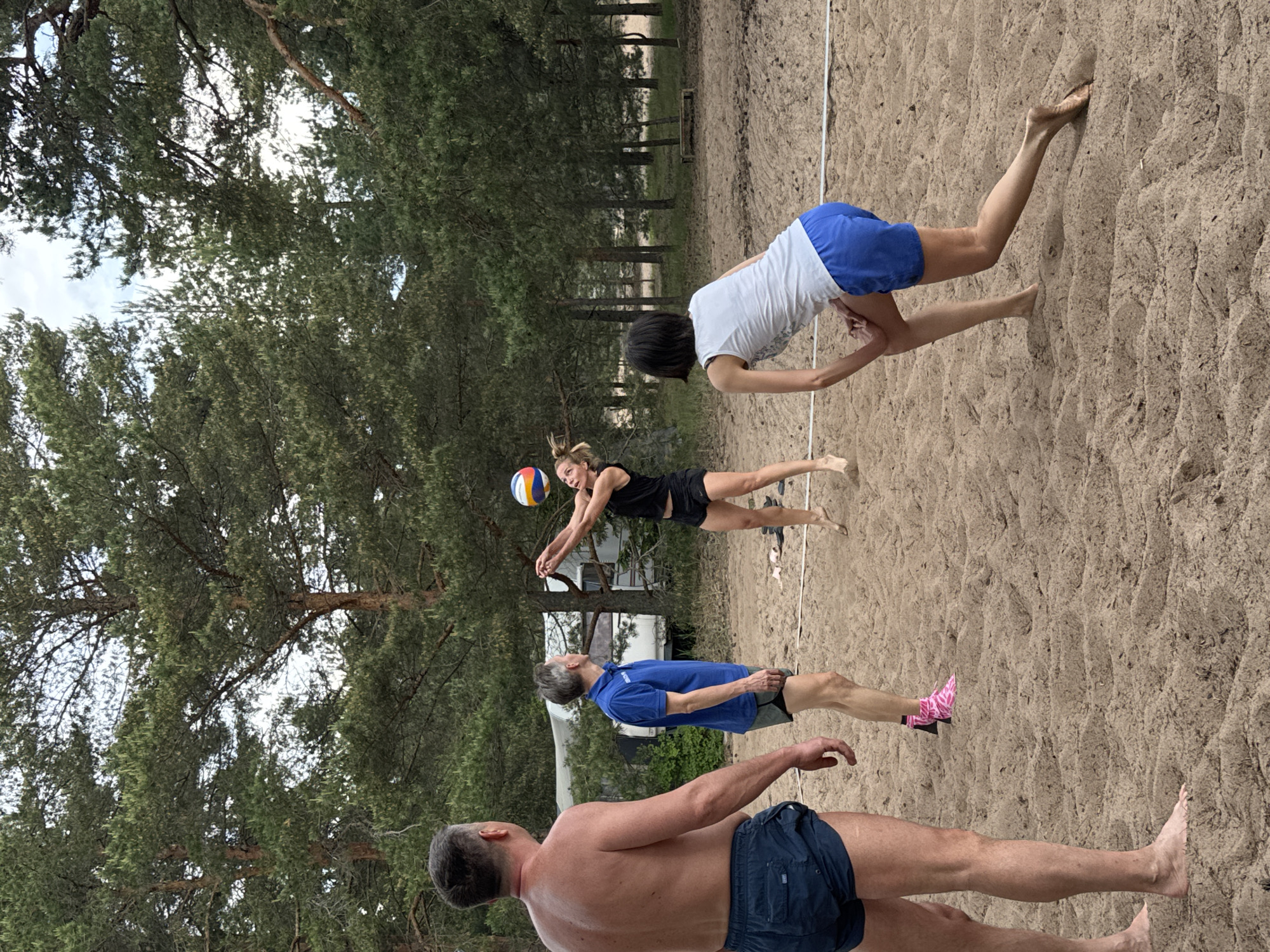 This image depicts an outdoor beach volleyball game taking place on a sandy court in Zelenaya Roshcha, Russia. Four adults are visible within the frame, engaged in or observing the play.
In the left foreground, an adult male with short grey hair stands with his back to the viewer. He is shirtless, wearing dark blue swim shorts, and is barefoot on the sand. His gaze appears directed towards the center of the court.
To his right, positioned further into the court, an adult male with short grey hair stands facing right. He wears a blue short-sleeved polo shirt and dark grey shorts. Pink patterned socks are visible on his feet, which are otherwise barefoot on the sand. He is looking towards the female player about to strike the ball.
In the middle-right of the frame, an adult female with blonde hair, pulled back, is captured mid-air. She wears a black tank top and black athletic shorts and is barefoot. She is in the process of hitting a multi-colored volleyball (orange, blue, white) with both hands, positioned above her head.
In the right foreground, an adult female with short dark hair stands in a crouched, ready position, facing the net and the opposite side of the court. She wears a white tank top and blue athletic shorts and is barefoot. Her left hand rests on her left knee.
A white line, indicating a boundary or the top/bottom of a net, stretches horizontally across the sand between the players. The ground is entirely light brown sand, marked with impressions and undulations.
The court is surrounded by a dense natural environment dominated by tall pine trees with dark green needles and brown trunks. In the background, partially obscured by trees and located behind the male in the blue shirt, two white recreational vehicles, resembling trailers or campers, are visible. Further to the right in the background, a simple wooden bench is situated at the edge of the sandy area. The sky, visible through the tree canopy, appears overcast.