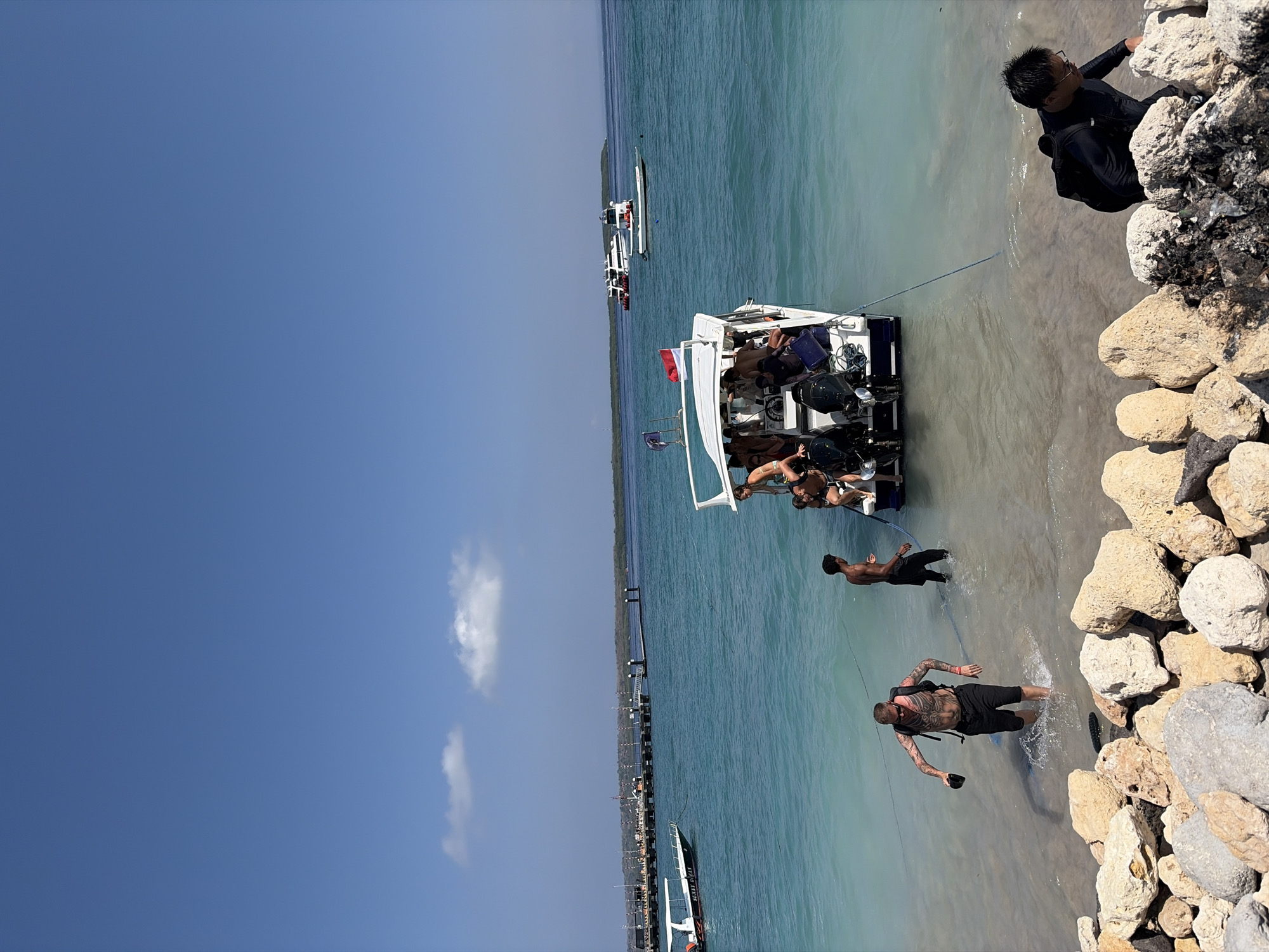 The image depicts a coastal scene in Nusa Penida, Indonesia, under a clear blue sky with scattered white clouds. The foreground is occupied by a rocky shoreline. In the middle ground, a white speed boat with a canopy and an Indonesian flag is partially submerged in shallow, turquoise water. Several people are either boarding or disembarking the boat. One man, heavily tattooed and wearing black shorts and a backpack, is wading in the water towards the shore, holding a black object in his hand. Another man in dark clothing stands in the water near the boat. The boat itself is crowded with passengers. In the background, a long wooden pier extends into the ocean, with several other boats docked or moving in the distance. The water transitions from a very light, almost transparent shade near the shore to a deeper blue further out. The overall impression is a busy tropical location, likely a departure or arrival point for water activities.