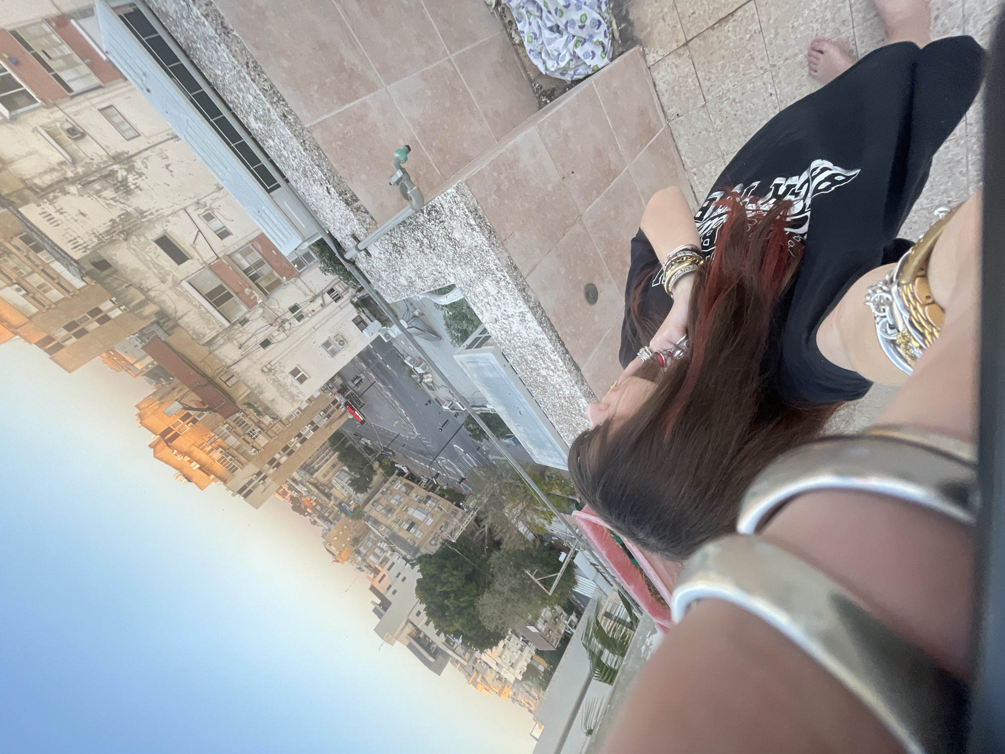 A high-angle shot shows a person standing on a roof in Petah Tikva, Israel, with a view of the city's buildings and streets in the background. The person is wearing black pants and a black t-shirt with a white design. They have brown hair with streaks of red and are wearing multiple bracelets on their wrist. The roof is made of beige tiles, and there is a water faucet visible near the edge. The sky is blue, and the light suggests it may be morning or late afternoon.