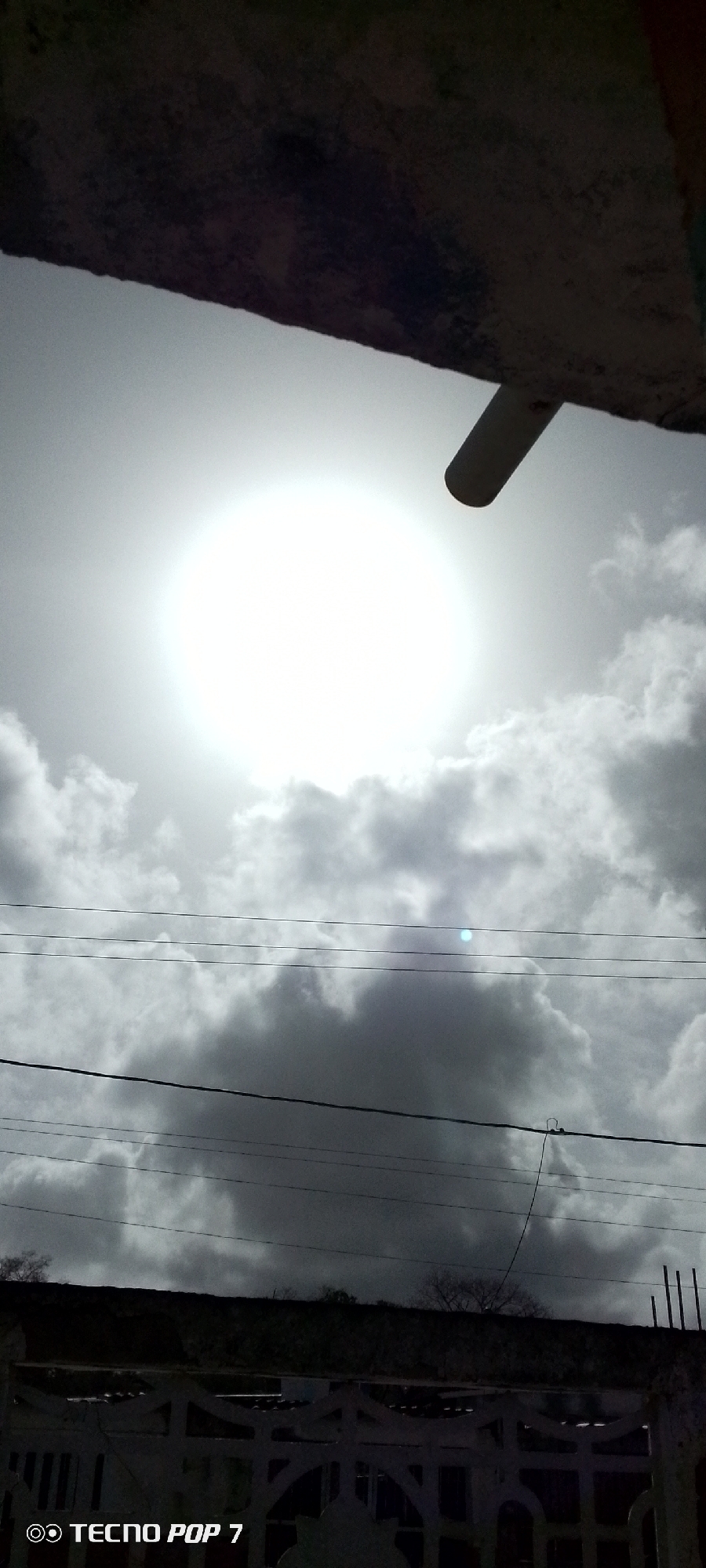 The image, taken in Altagracia, Venezuela, depicts a view of the sky obscured by clouds. A bright sun is visible through the cloud cover. Foreground elements include a partial view of a structure or building at the top of the frame and the presence of power lines bisecting the scene horizontally. A fence-like structure is at the bottom. The photo appears to have been taken from ground level, looking upwards.