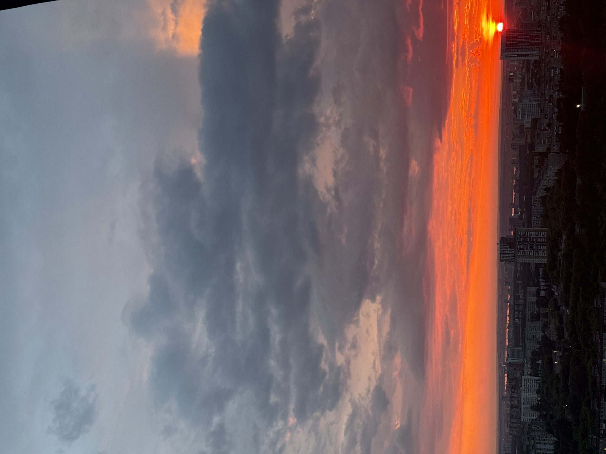 The image depicts a sunset over the city of Savenki, Russia. The sky is dominated by vibrant hues of orange and red, with dark clouds scattered throughout. The sun, a brilliant ball of light, is partially obscured by a building on the right side of the frame. Below the sky, a cityscape stretches across the horizon, featuring a mix of residential buildings and taller structures. Trees and greenery are visible in the foreground, suggesting a park or wooded area within the city. The overall scene is one of a dramatic and colorful end to the day.