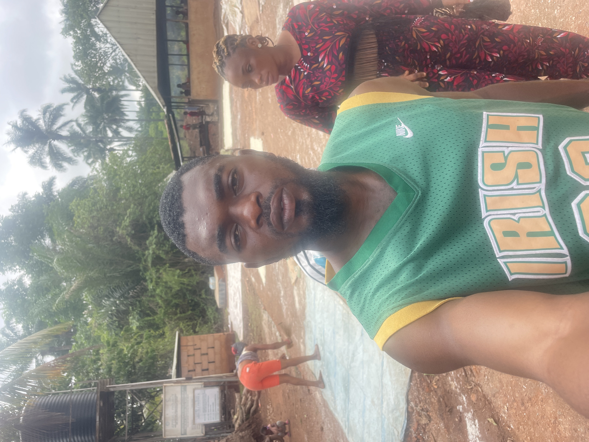 The image shows a man in a green and yellow "IRISH" jersey taking a selfie with a woman in a red patterned shirt in the background. The background indicates a rural or semi-rural setting with trees, a building with a visible roof, and other people, including a person in orange shorts walking. The location is Eke, Nigeria.