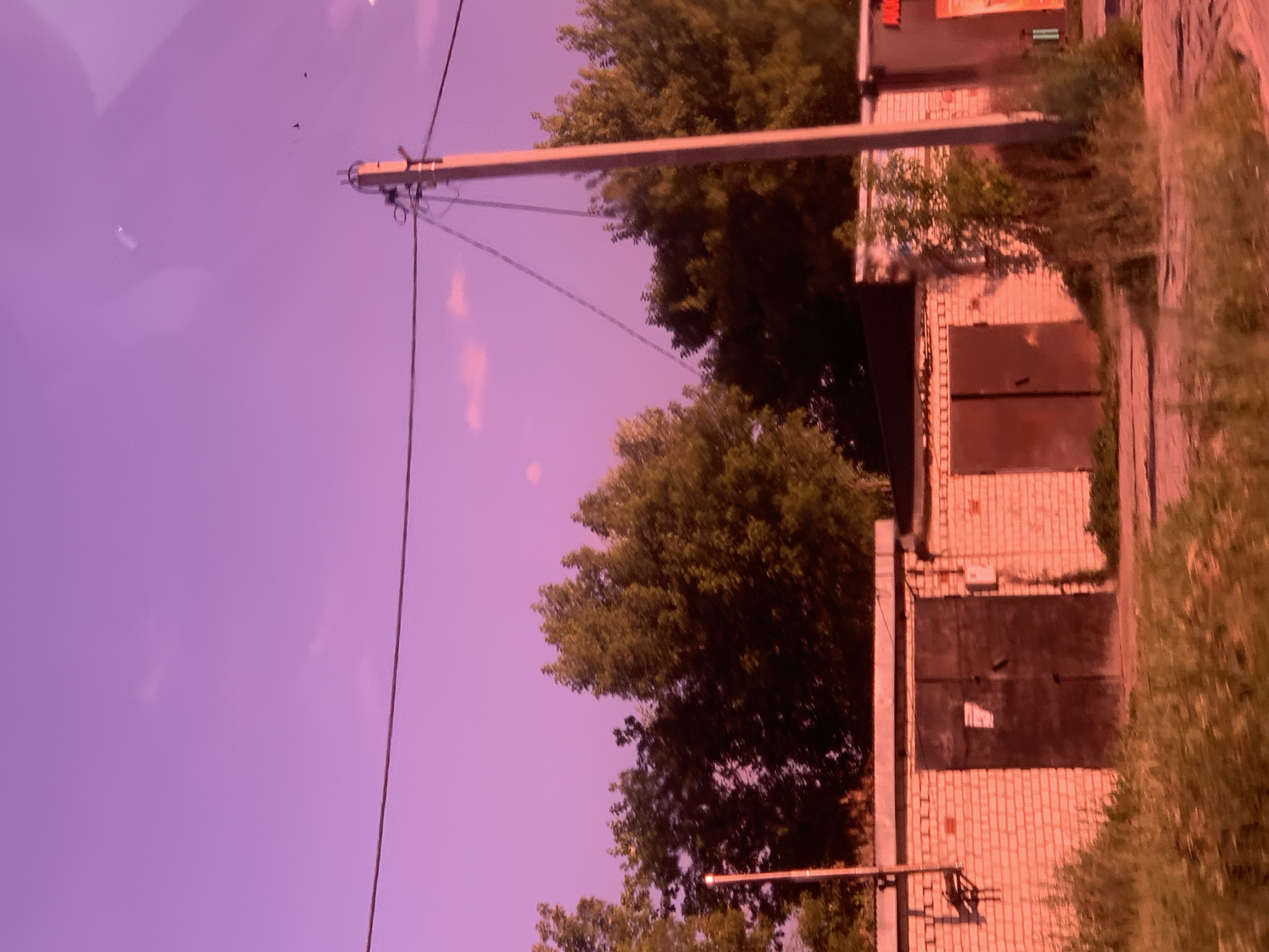 The image depicts a row of garages constructed from white brick, set against a backdrop of lush green trees. The sky is a vibrant purple, suggesting either a sunset or sunrise. In the foreground, dry grass and vegetation line the ground in front of the garages. A telephone pole stands to the right of the garages, with wires extending across the frame. The overall scene appears to be an outdoor urban environment, possibly in the city of Pavlovo, Russia, given the description provided. No people or specific actions are visible in the image.